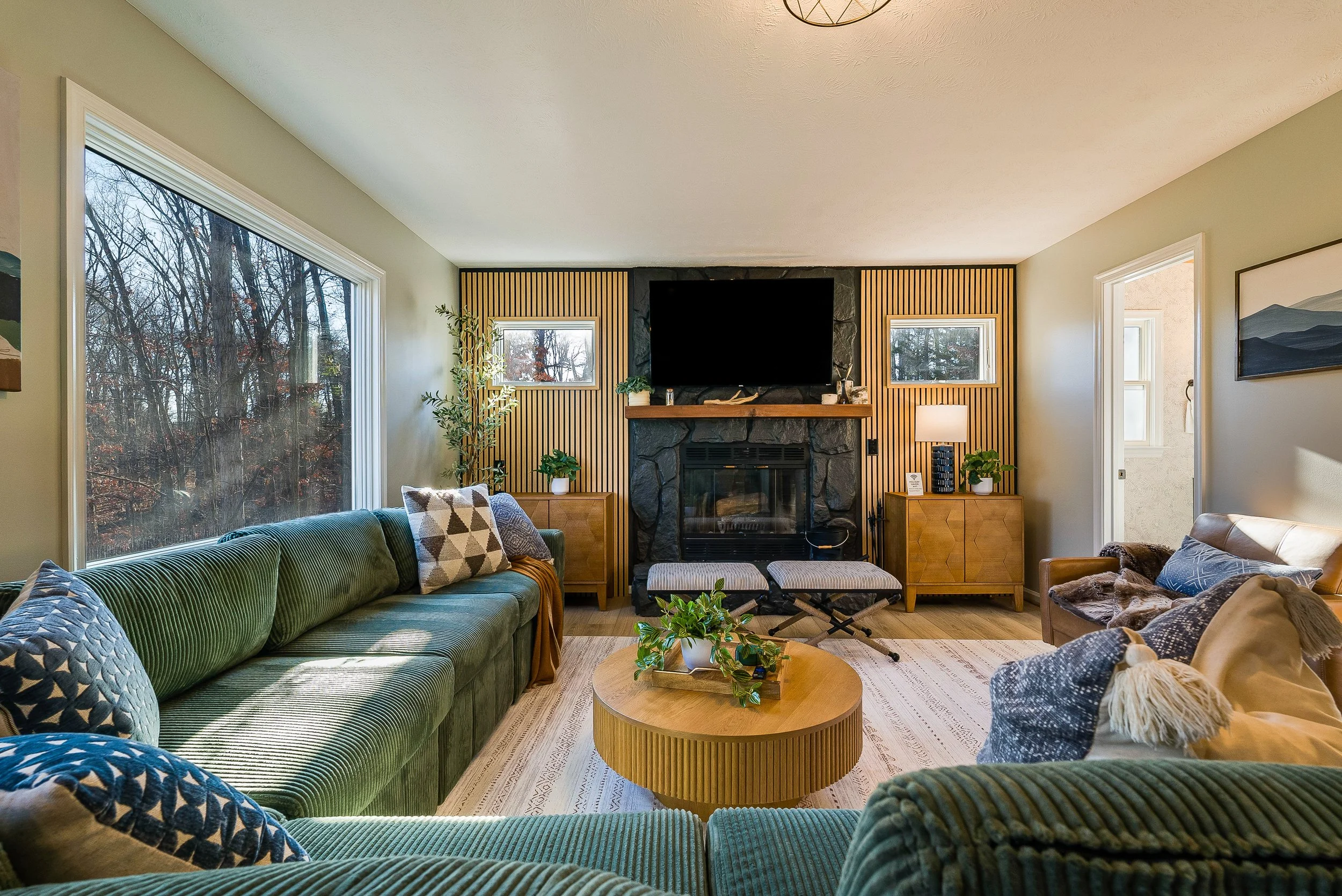 Massanutten Airbnb interior design of a living room with windows showing, a wood-paneled accent wall with a black painted stone fireplace, green velvet sofa with patterned pillows, a wooden coffee table and side tables with lamps and plants.
