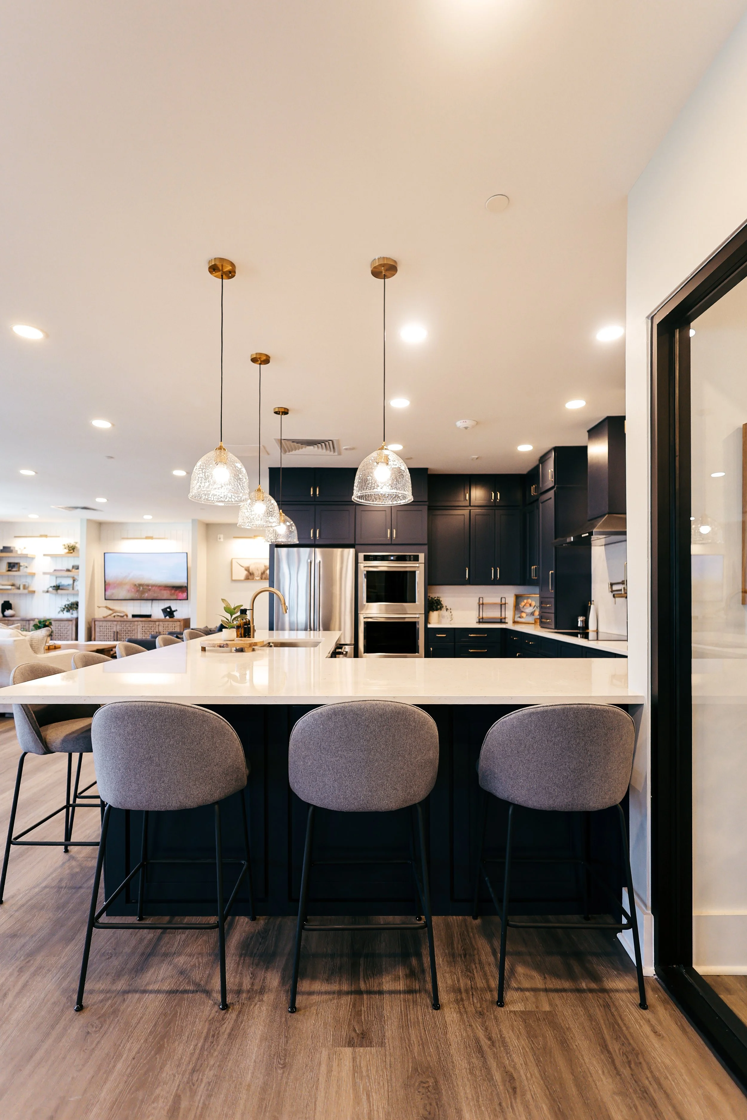 Modern kitchen with dark cabinetry, a large island with beige counter, pendant lights, stainless steel refrigerator, double oven, and bar stools.