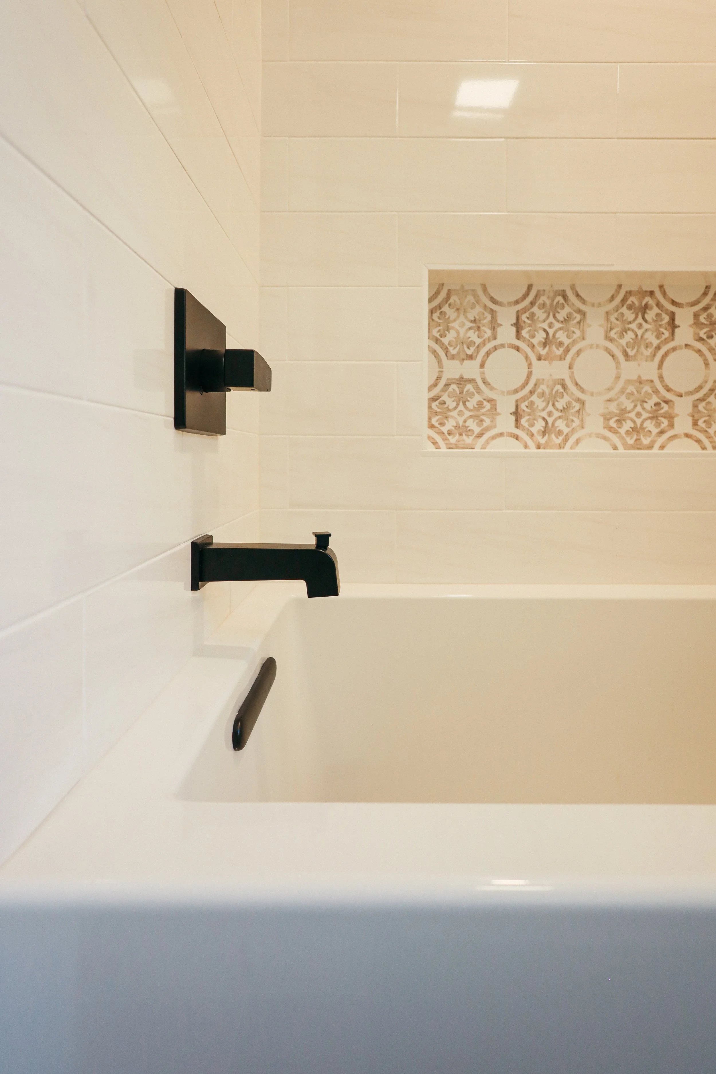 Close-up of a bathtub faucet and handle in a modern bathroom with beige tiles and decorative accent tiles behind the tub.