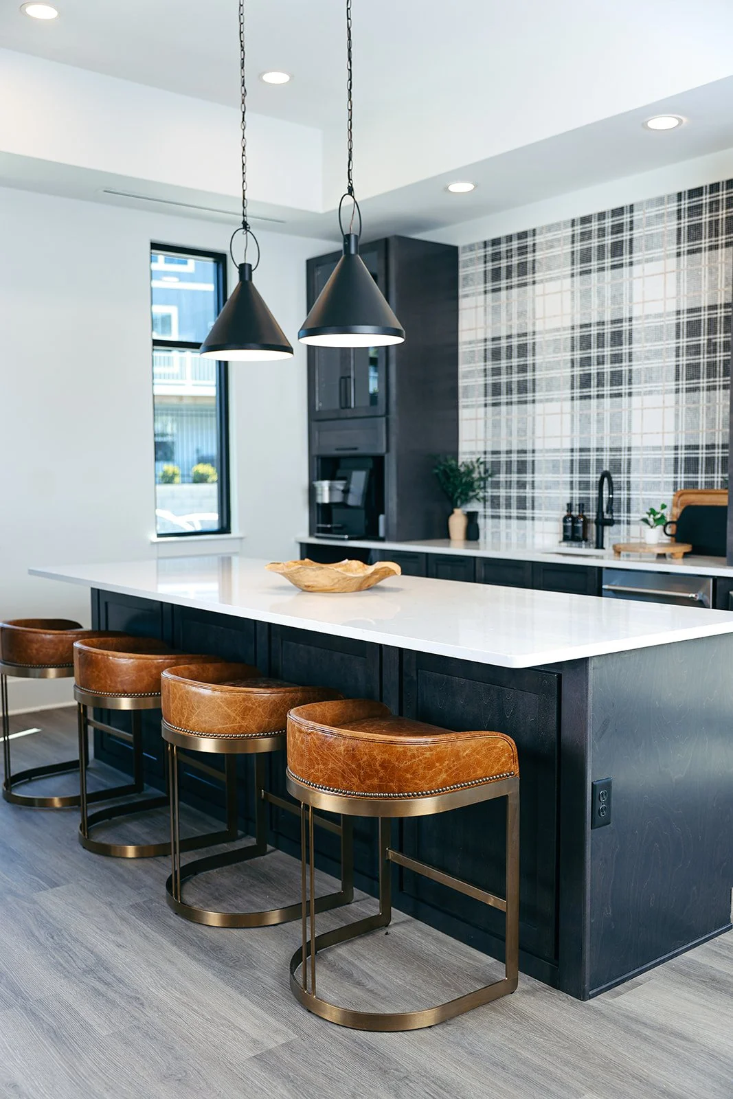Modern kitchen with black cabinets, white marble island, leather bar stools, pendant lighting, and patterned backsplash.