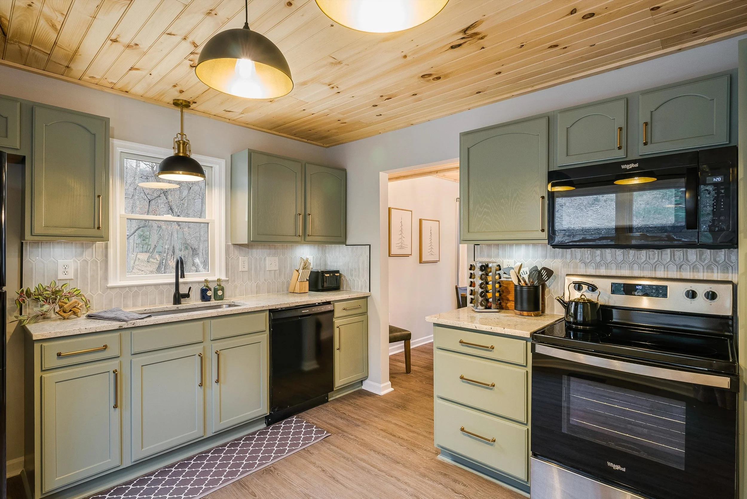 The image shows a modern kitchen with green cabinets and brass handles, a black stove and microwave, a granite countertop, and a wood-paneled ceiling. There are pendant lights hanging over the sink and a small window above the sink, with a view of trees outside. The kitchen also features a patterned rug on the wooden floor.