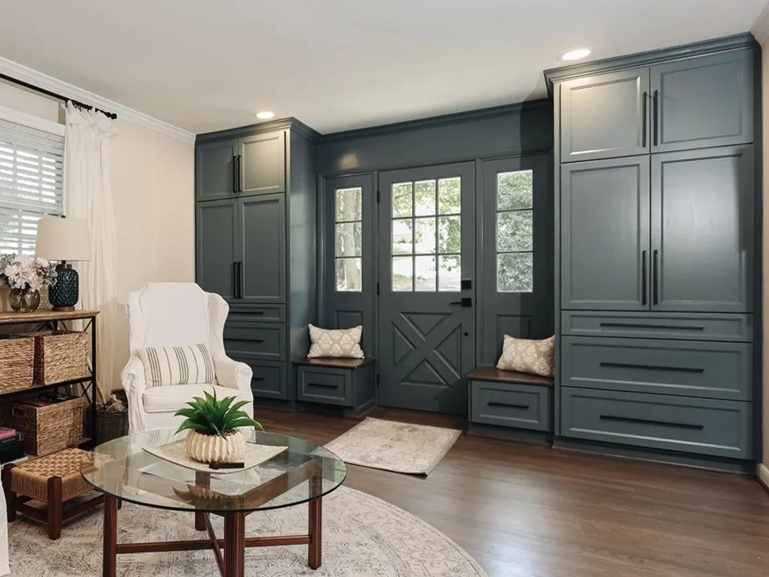 Modern kitchen with white cabinets, stainless steel appliances, hardwood floors, and decorative rugs, leading to a living room with a sofa and curtains.
