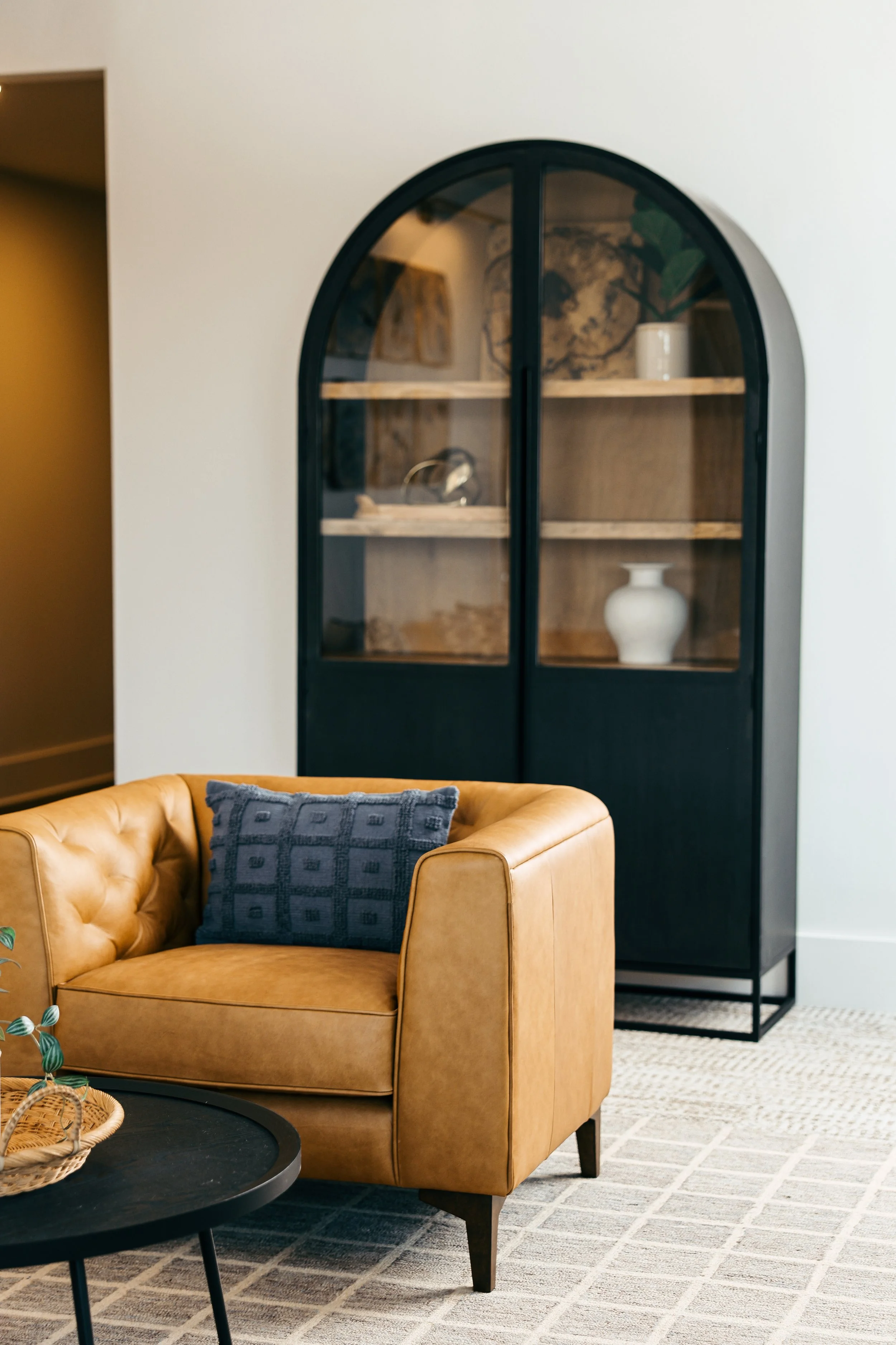 Office seating area with a caramel leather sofa, a navy blue cushion, a black round coffee table, a beige and white patterned rug, and a black display cabinet with glass doors holding decorative items.