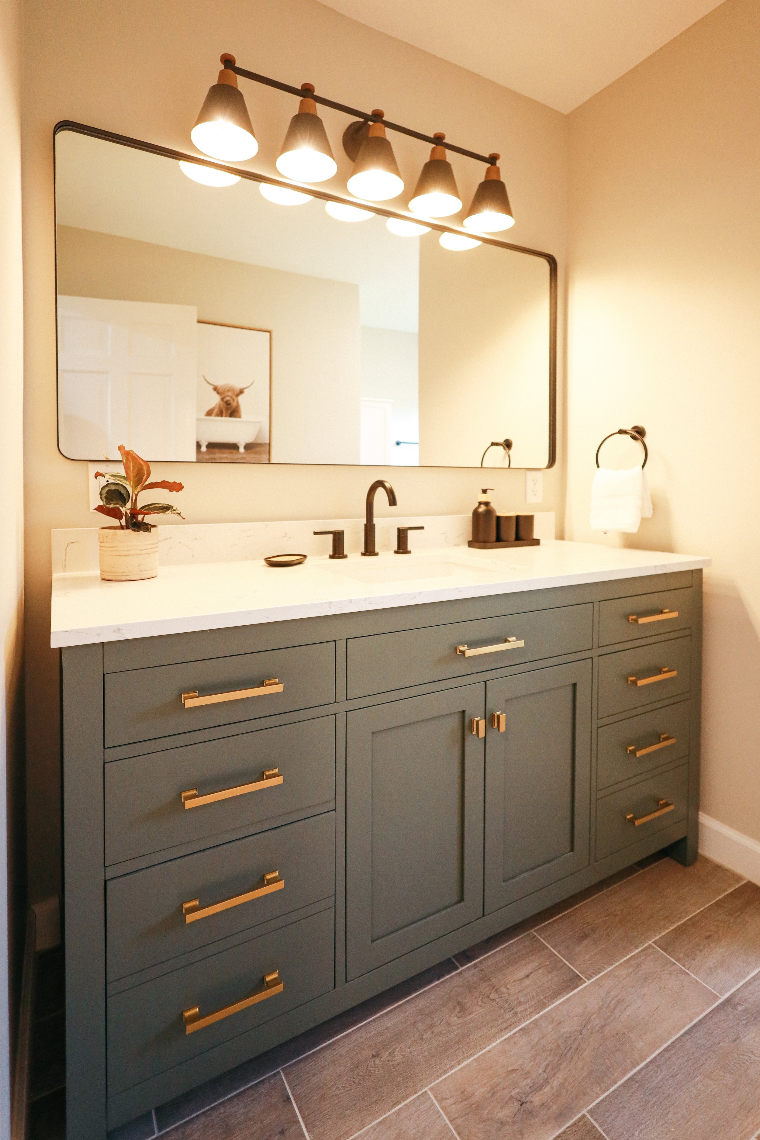 Bathroom vanity with a large mirror, six gold handles, a black faucet, potted plant, soap dispenser, and towel ring.