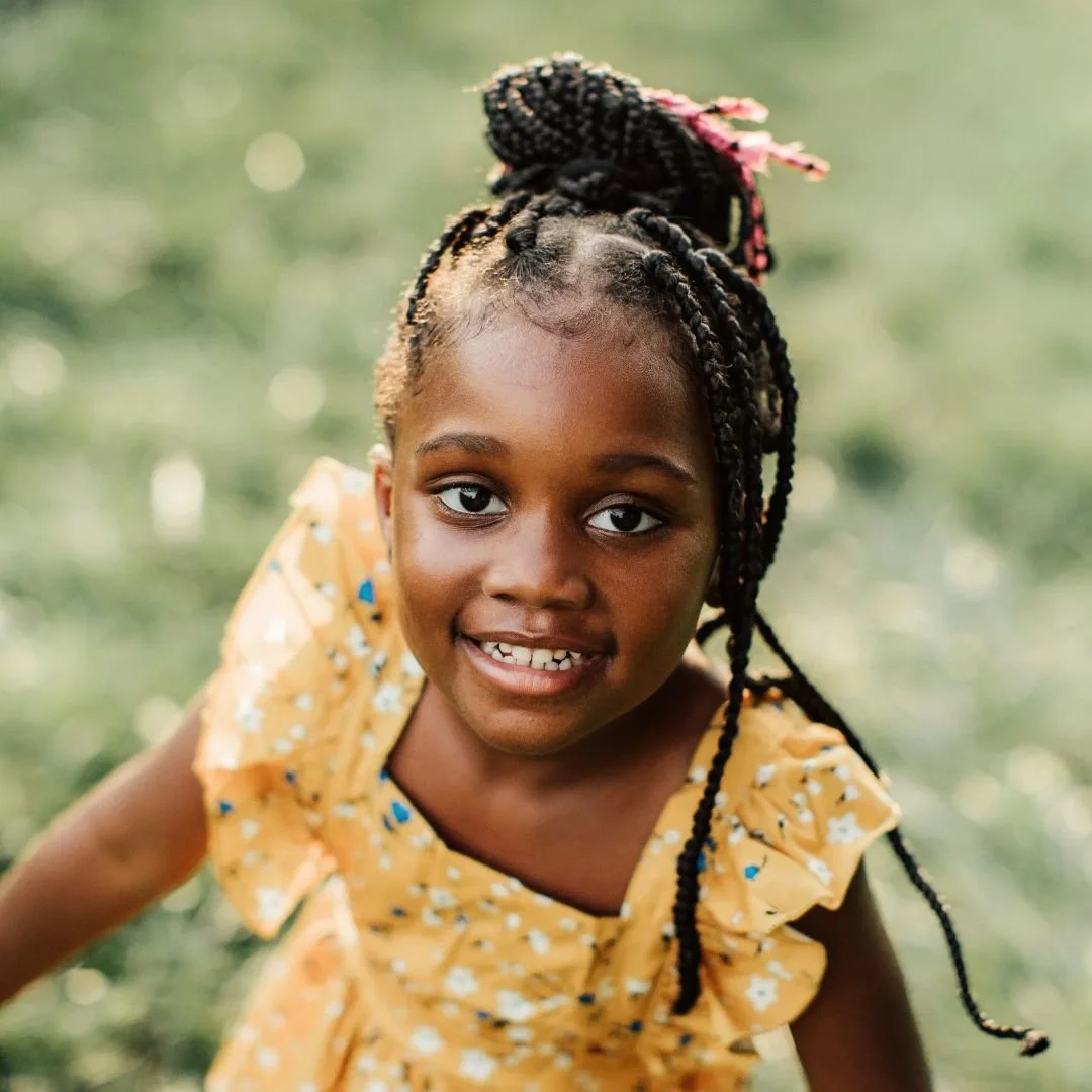 A close-up shot of a happy young Black girl with braided hair and a yellow floral dress smiling at the camera outdoors on a grassy field.