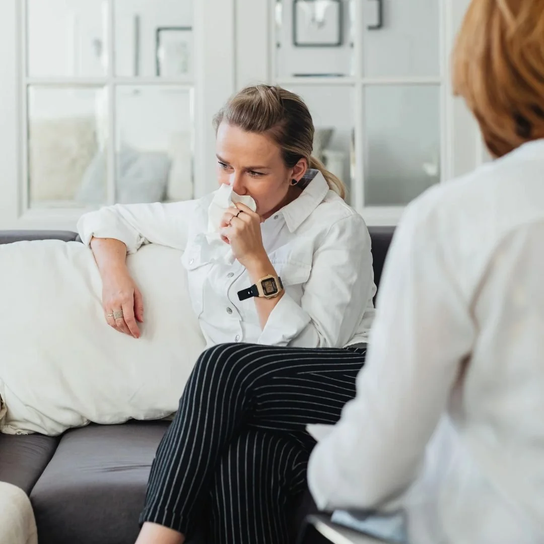A crying woman sitting on a couch holding a tissue, speaking to a therapist during a psychological trauma therapy session.