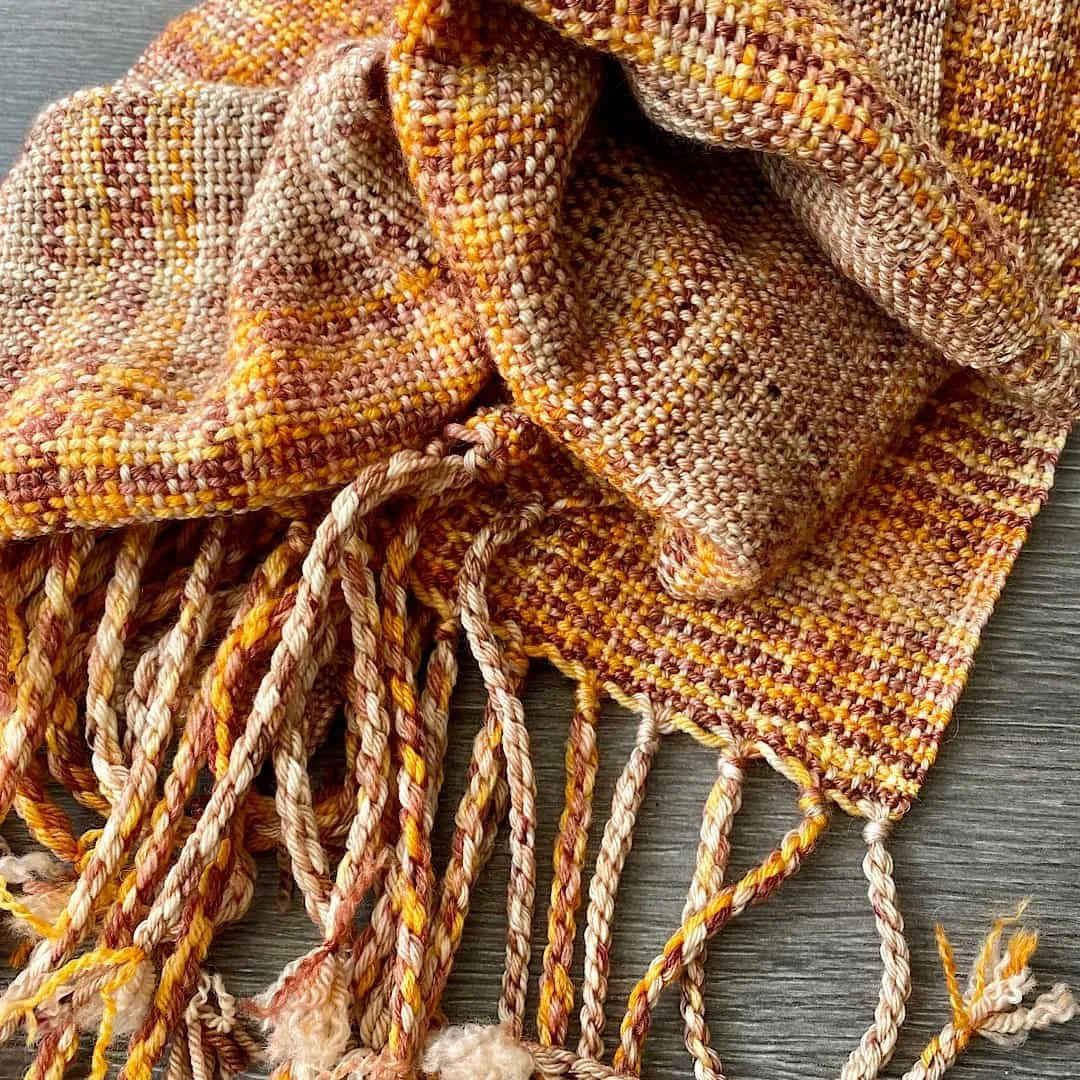 Ein handgewebter Schal mit Fransen in warmen Farben wie Gelb, Orange und Braun, auf einem Holzuntergrund liegend.