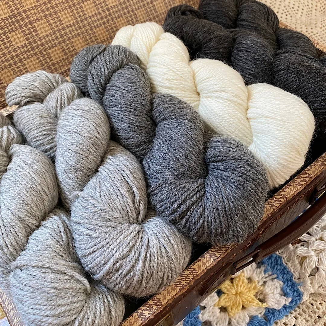 Mehrere Knäuel Wolle in verschiedenen Grautönen, Weiß und Schwarz, auf einem Korb mit einer türkis-blauen und weißen geflochtenen Decke darunter.