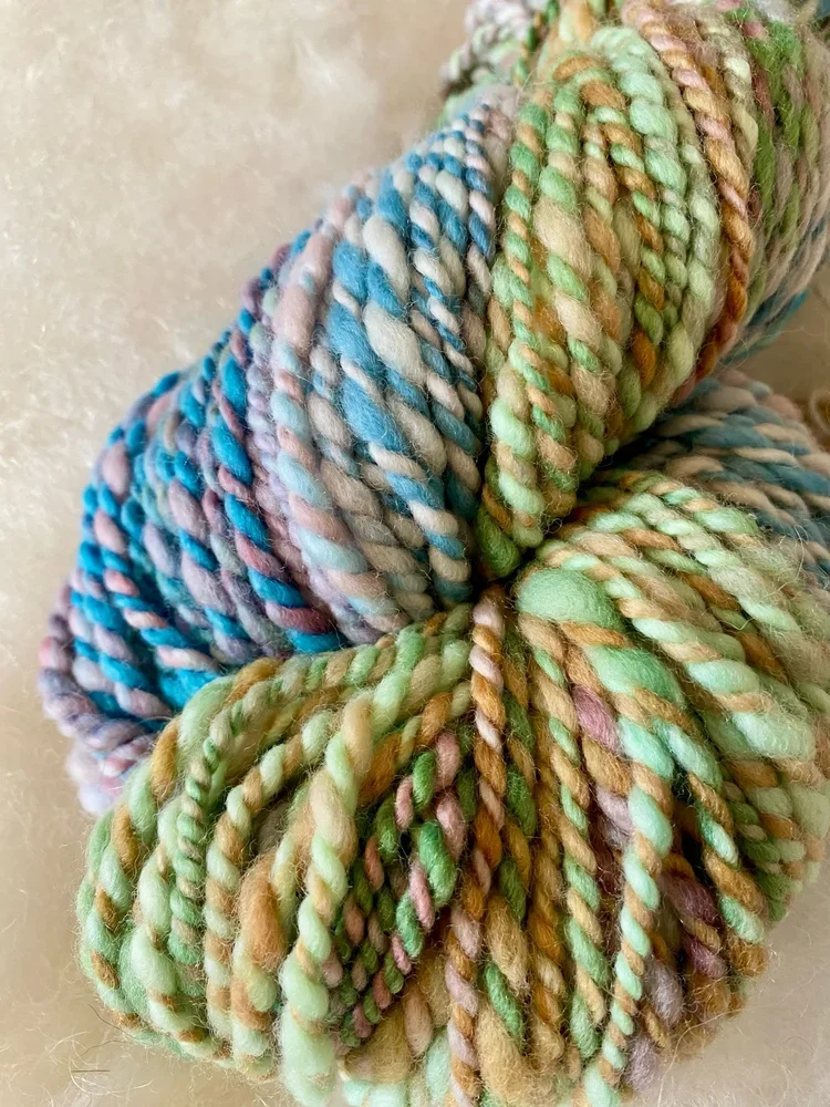 Dünne, bunte Wolle in Grüntönen, Blau, Violett und Braun, zu einem Knäuel aufgerollt.