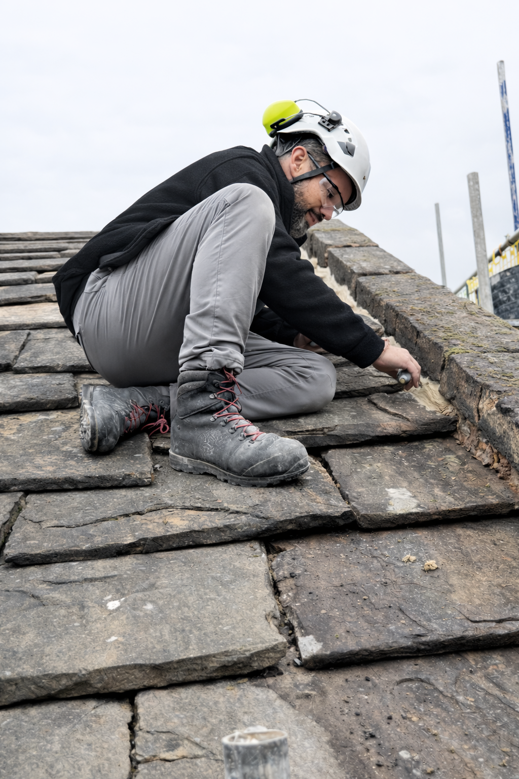 A man wearing a safety helmet, glasses, and work boots is kneeling on a stone roof, using a tool to work on the roofing material.