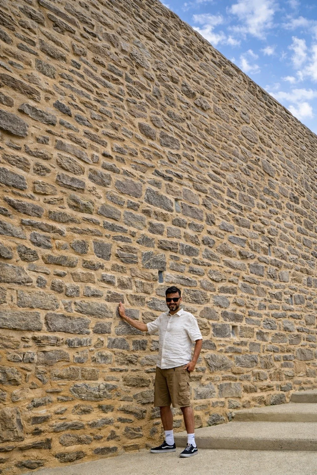 A man in a white shirt, beige shorts, and sneakers stands next to a large stone wall, touching it with his left hand, and wearing sunglasses, with a partly cloudy sky in the background.