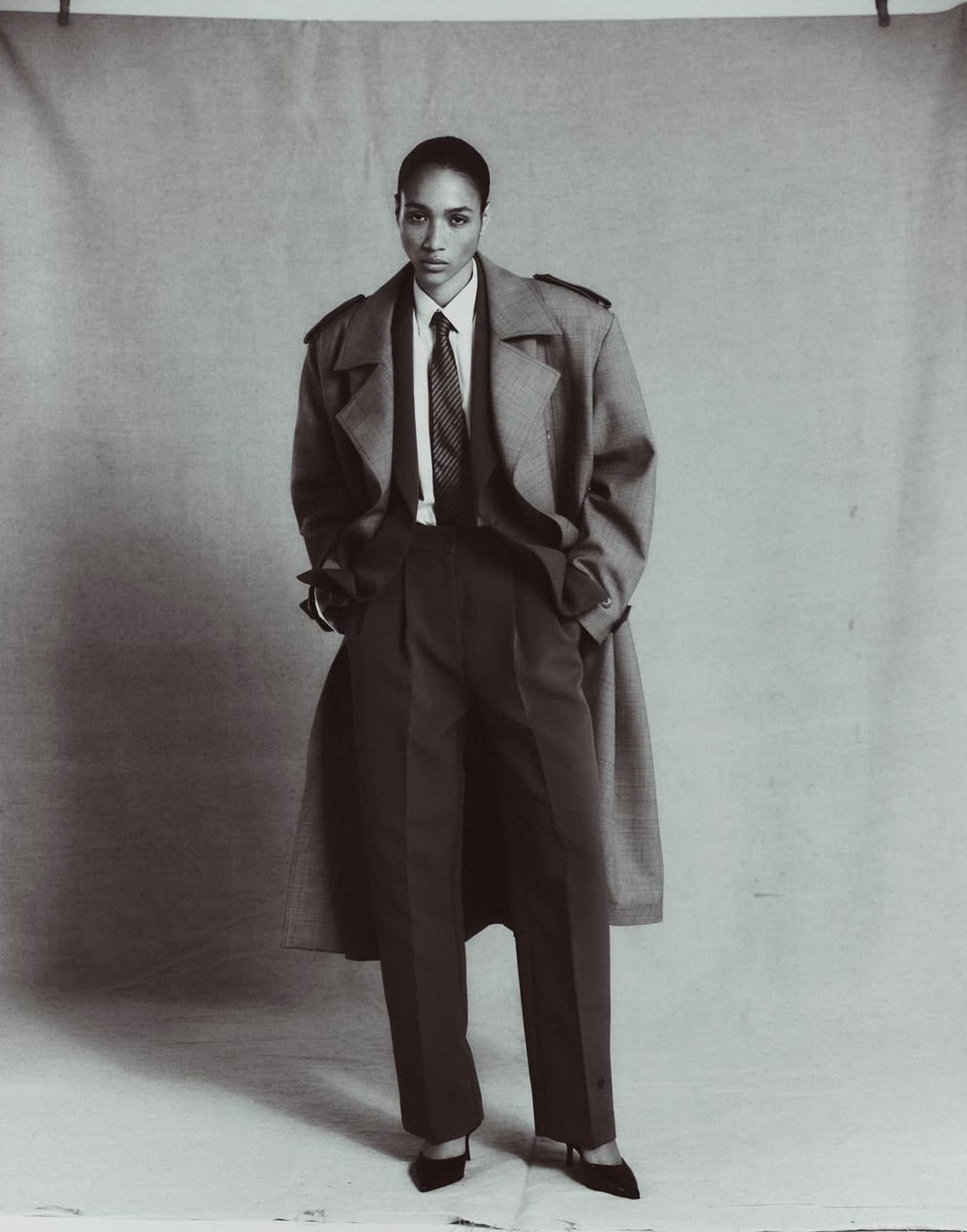 Une femme pose avec un manteau long, costume avec une chemise et une cravate, et des chaussures à talons, dans une image en noir et blanc.