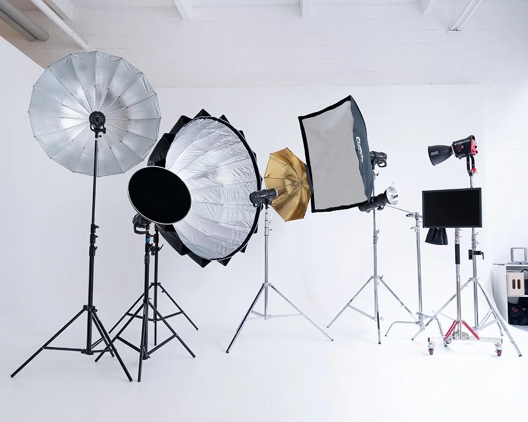 Studio photo avec éclairages et supports d'éclairage, notamment parapluies, softbox, projecteur et écran, sur un fond blanc.