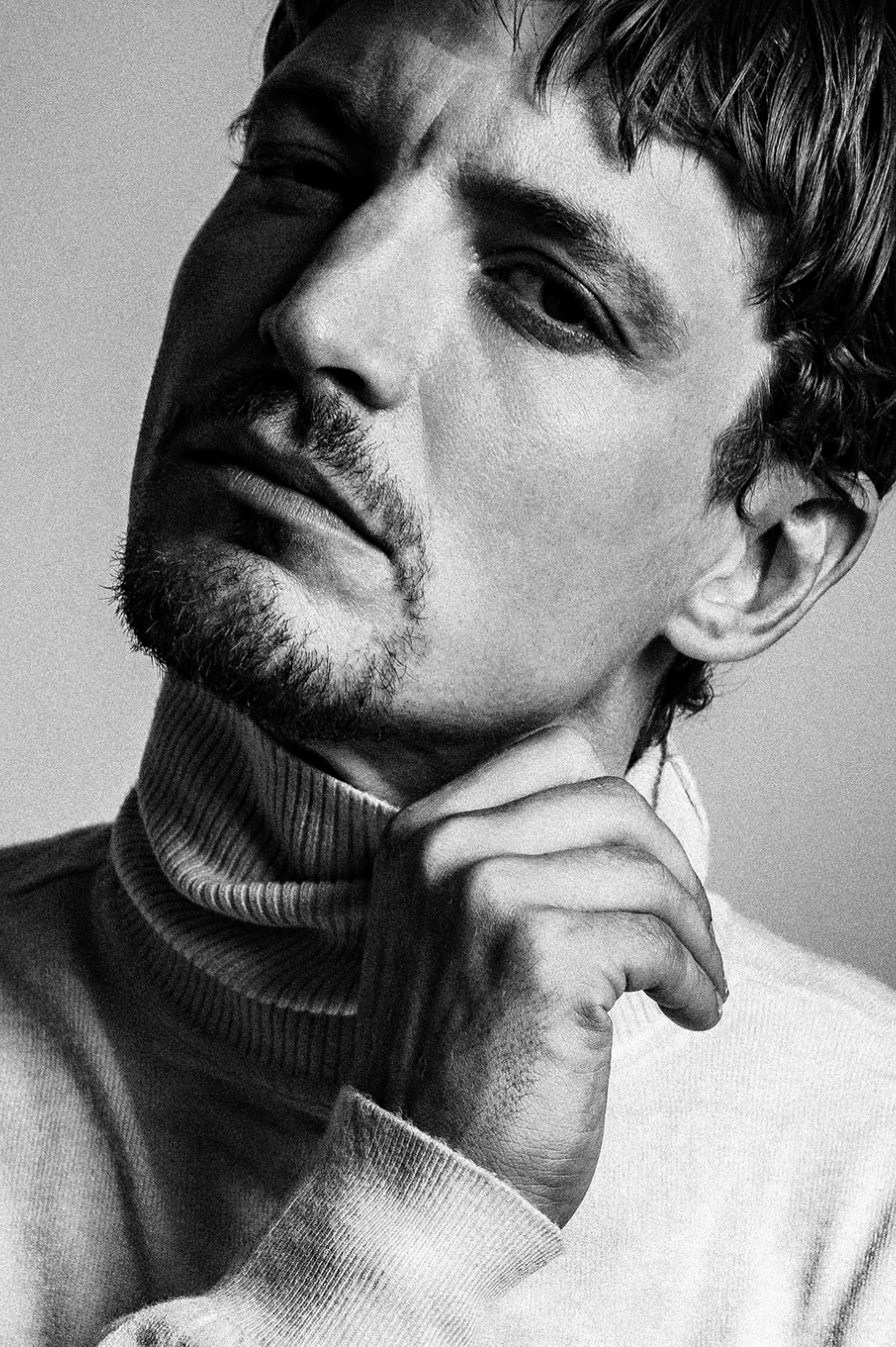 Neils Schneider. Portrait en noir et blanc d'un homme avec une main sous le menton, portant un col roulé.