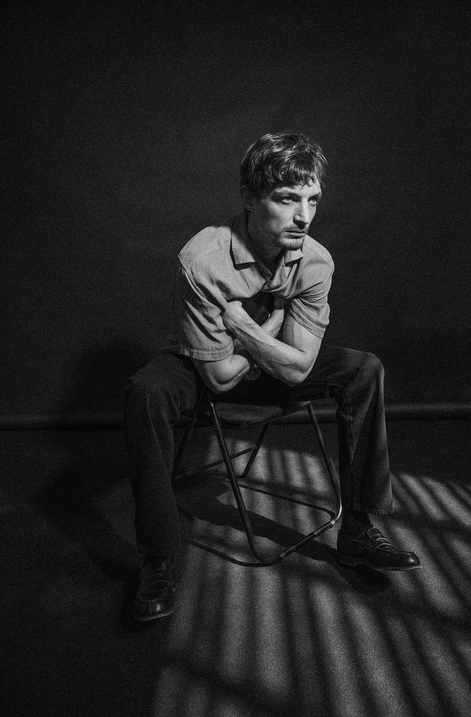Un homme assis sur une chaise dans une pièce sombre, éclairé par des lumières qui créent des ombres au sol. Neils Schneider