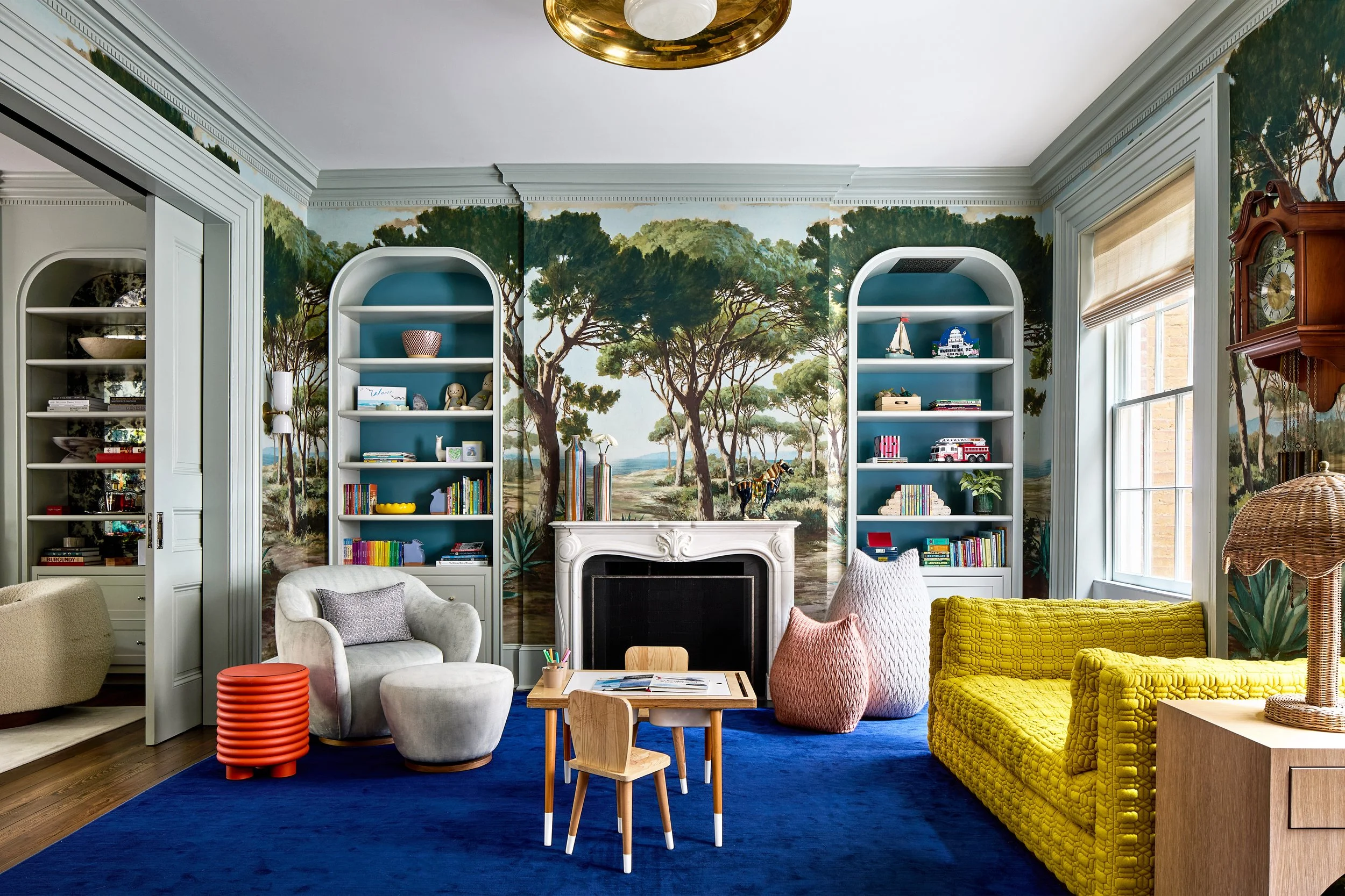 Living room with painted jungle scene wallpaper, white fireplace, colorful furniture including gray, pink, and yellow chairs, blue rug, and built-in bookshelves with books and decorative items.