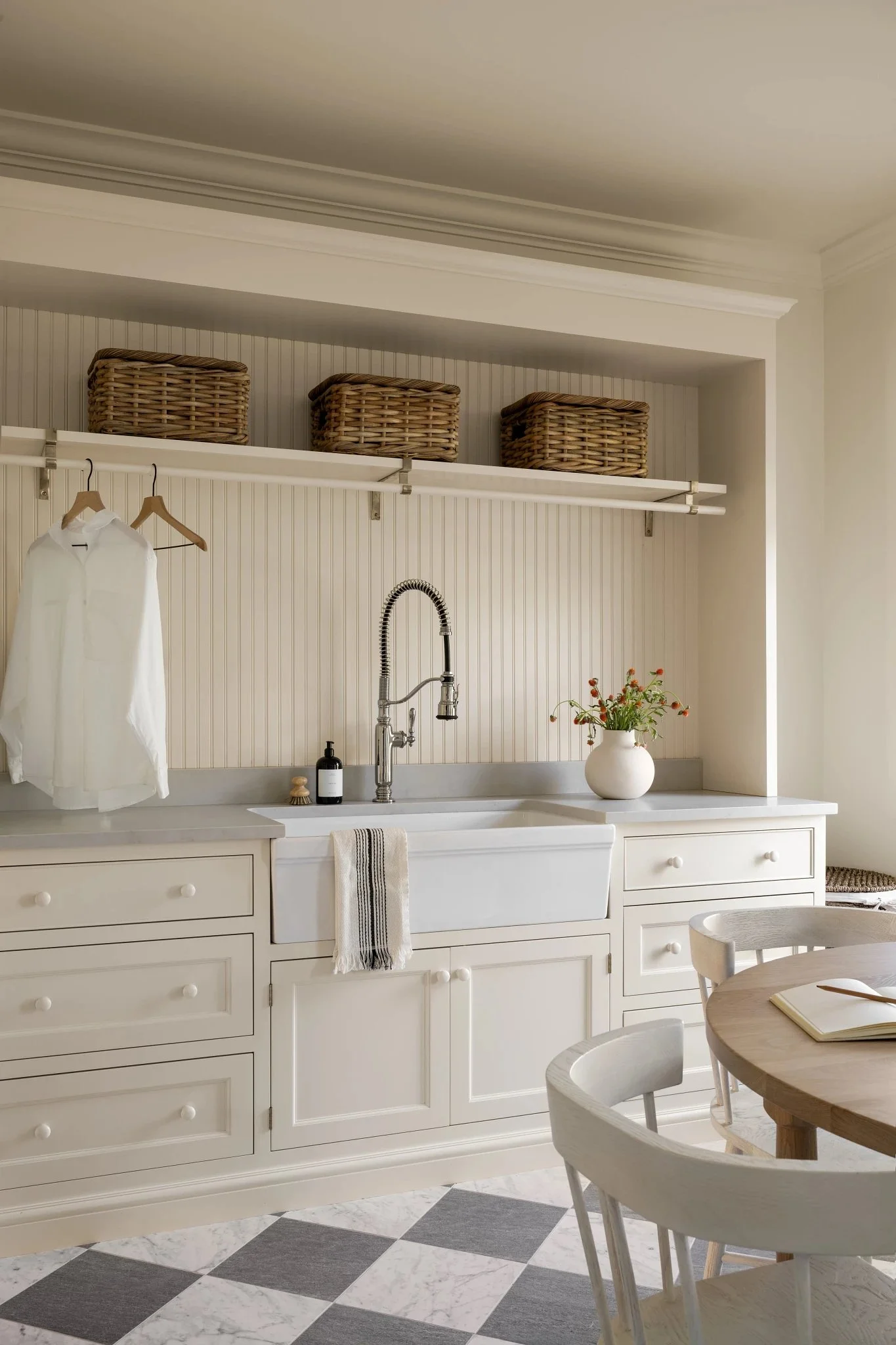 A cozy kitchen with white cabinets, a white farmhouse sink, and a checkered black and white tile floor. Wicker baskets are stored on a shelf above. A white shirt is hanging on a hook, and a vase with flowers is on the countertop.