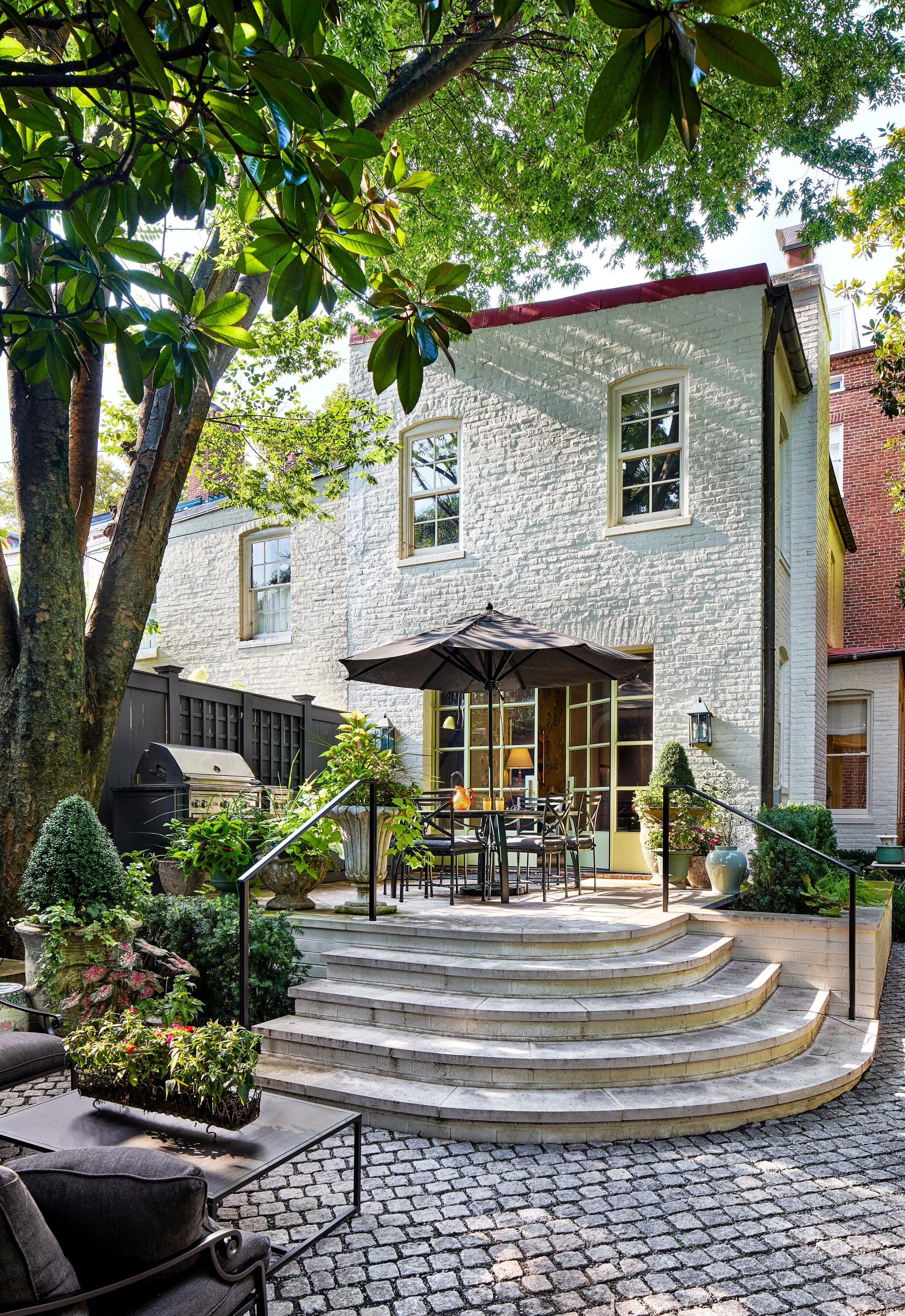 Backyard patio with a white brick house, potted plants, a round table with chairs and a large umbrella, surrounded by trees and greenery.