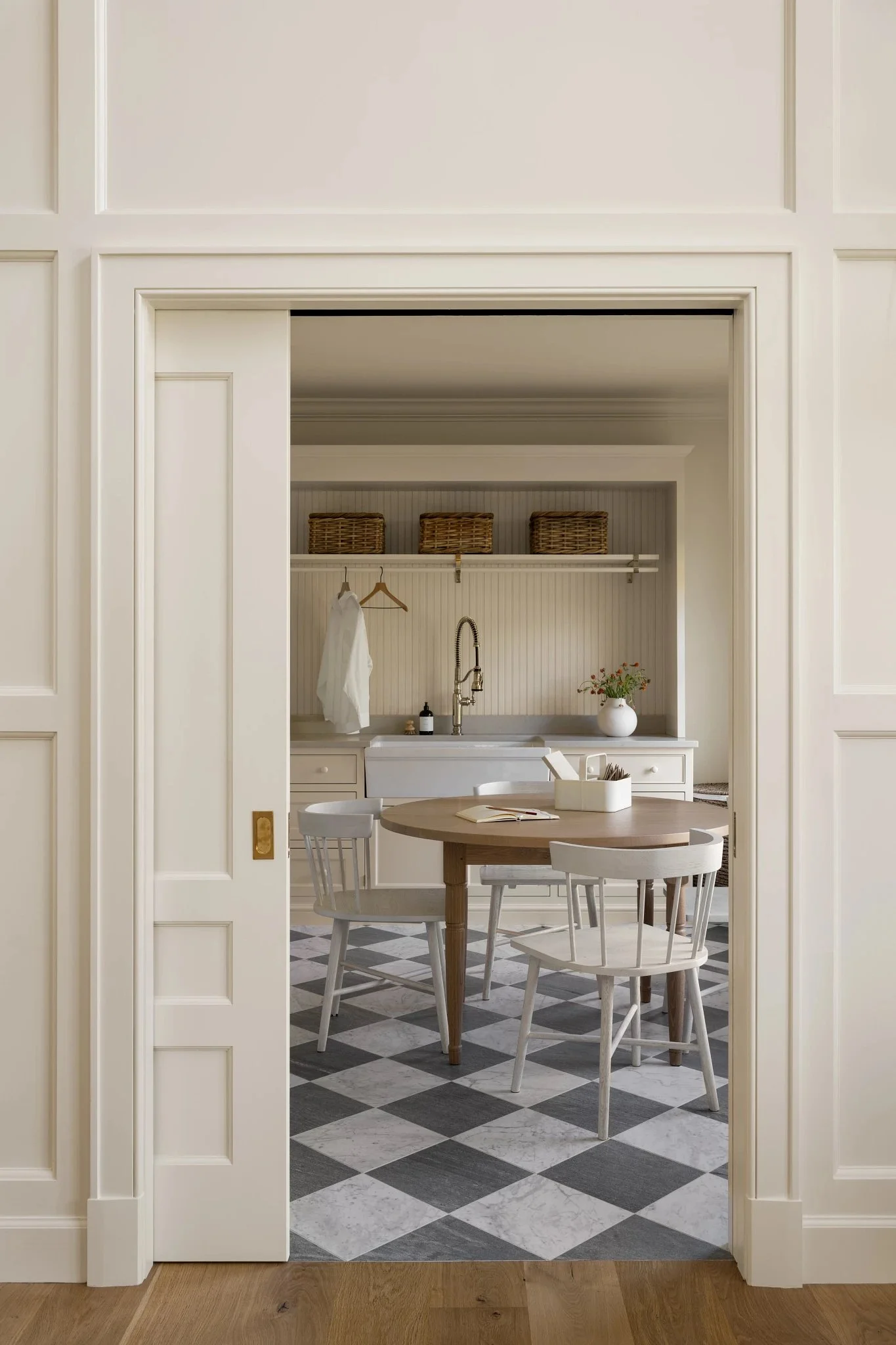 A view through a sliding door into a kitchen and dining area with a round wooden table and white chairs, a sink, hanging rod with a white towel, baskets on a shelf, and a checkered black and white tile floor.