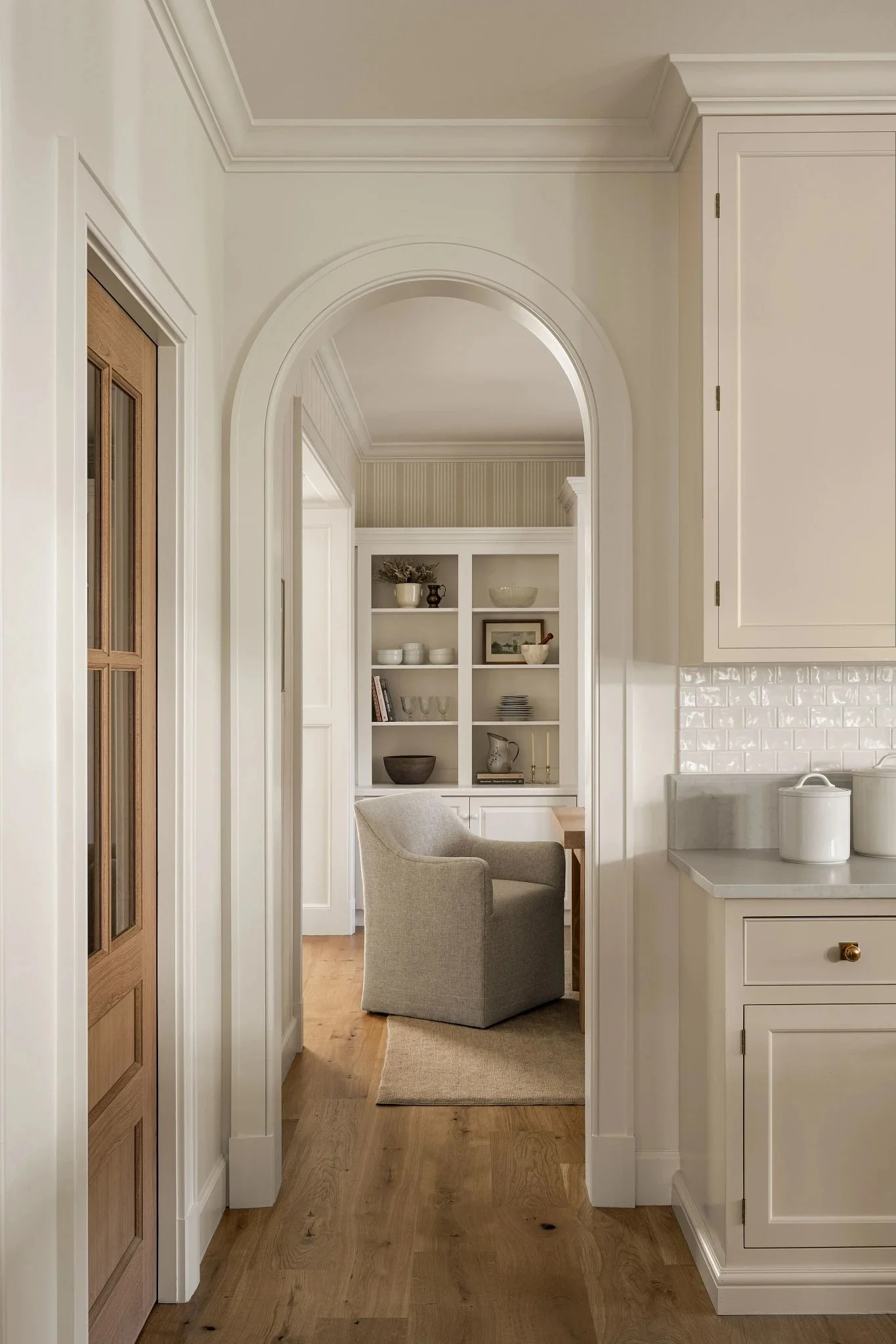 View of a cozy nook with a beige armchair, built-in shelves with decorative items and books, seen through an arched doorway from a kitchen area with white cabinets and a light wood floor.