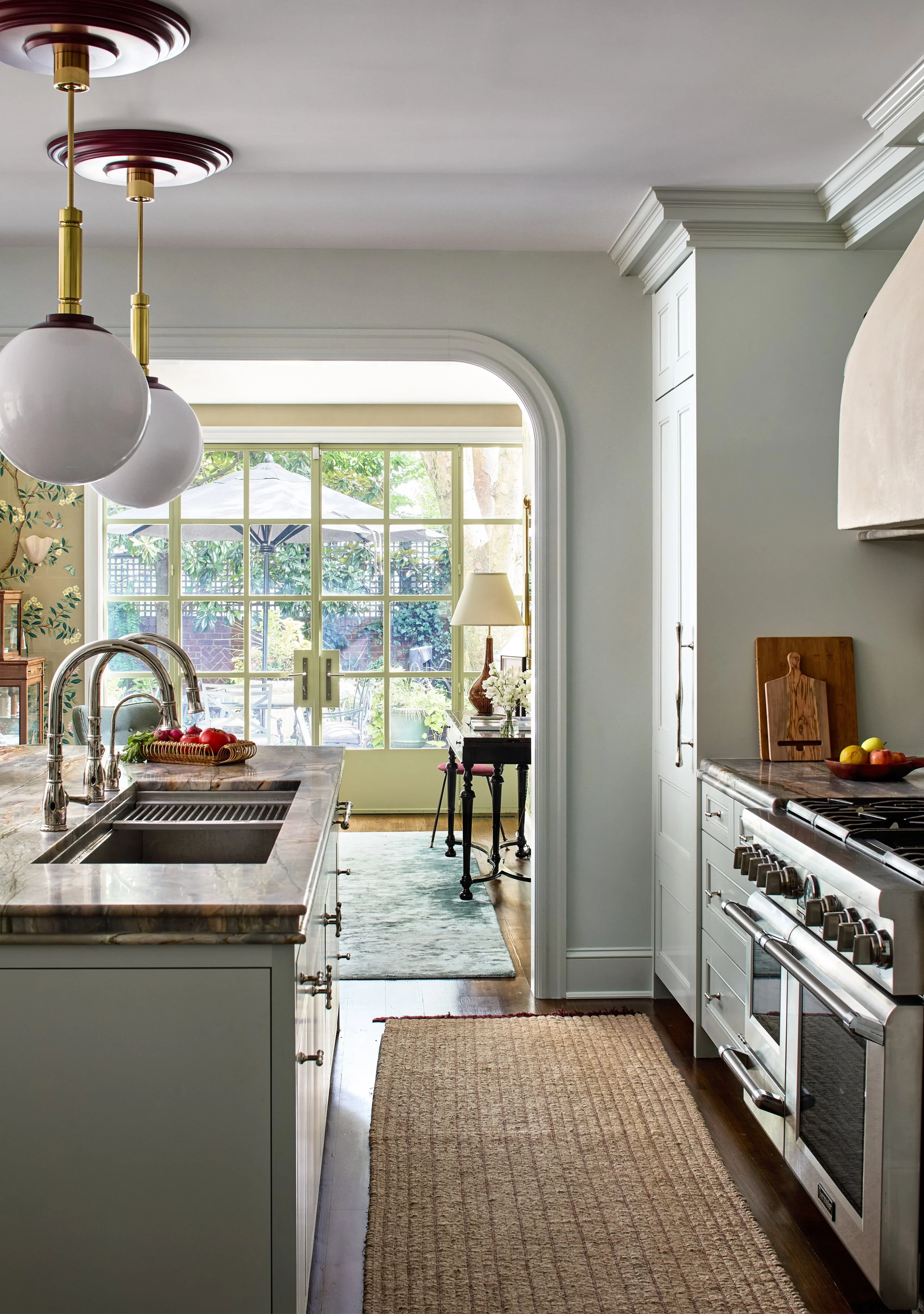 A bright, airy kitchen with white cabinetry, stainless steel appliances, a marble island with a sink, and a view to a sunlit patio garden through large glass doors.