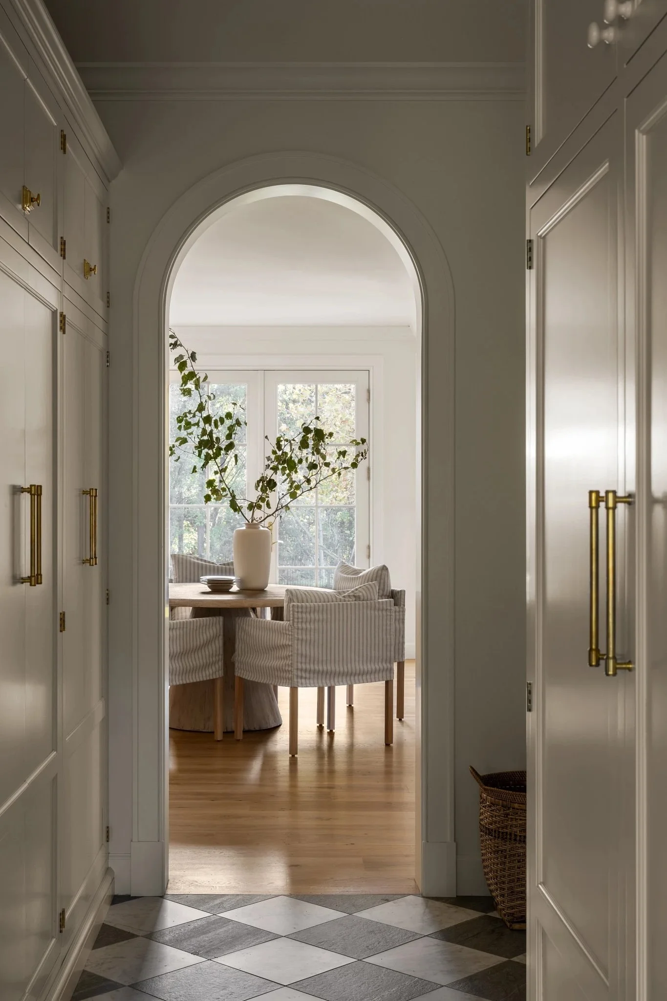 A view from a hallway into a dining room with a round table, striped upholstered chairs, a large white vase with leafy branches, and sunlight coming through a window