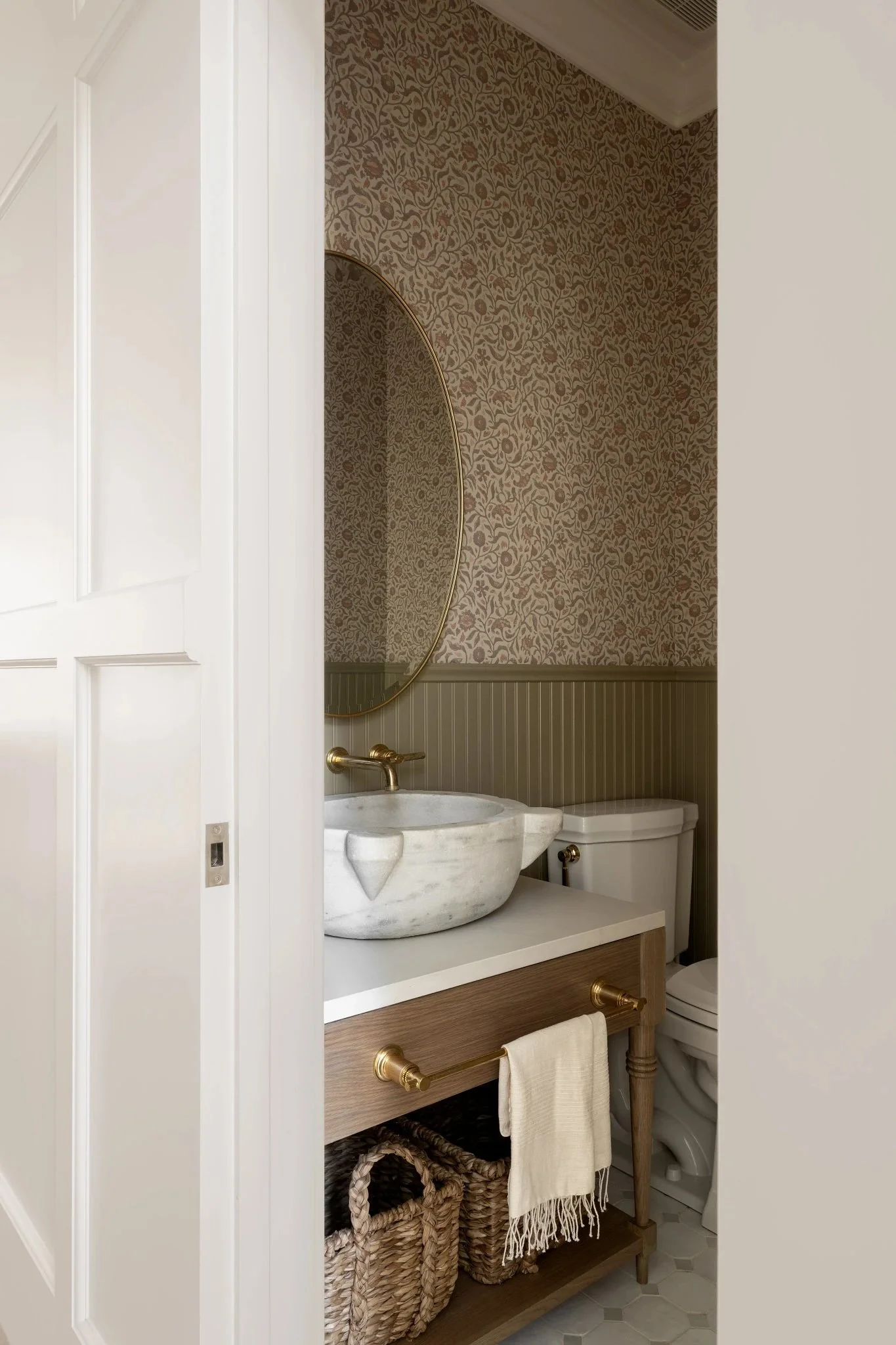 A small bathroom with a marble sink, a mirror, and a toilet. The wall has patterned wallpaper and there are woven baskets under the sink.