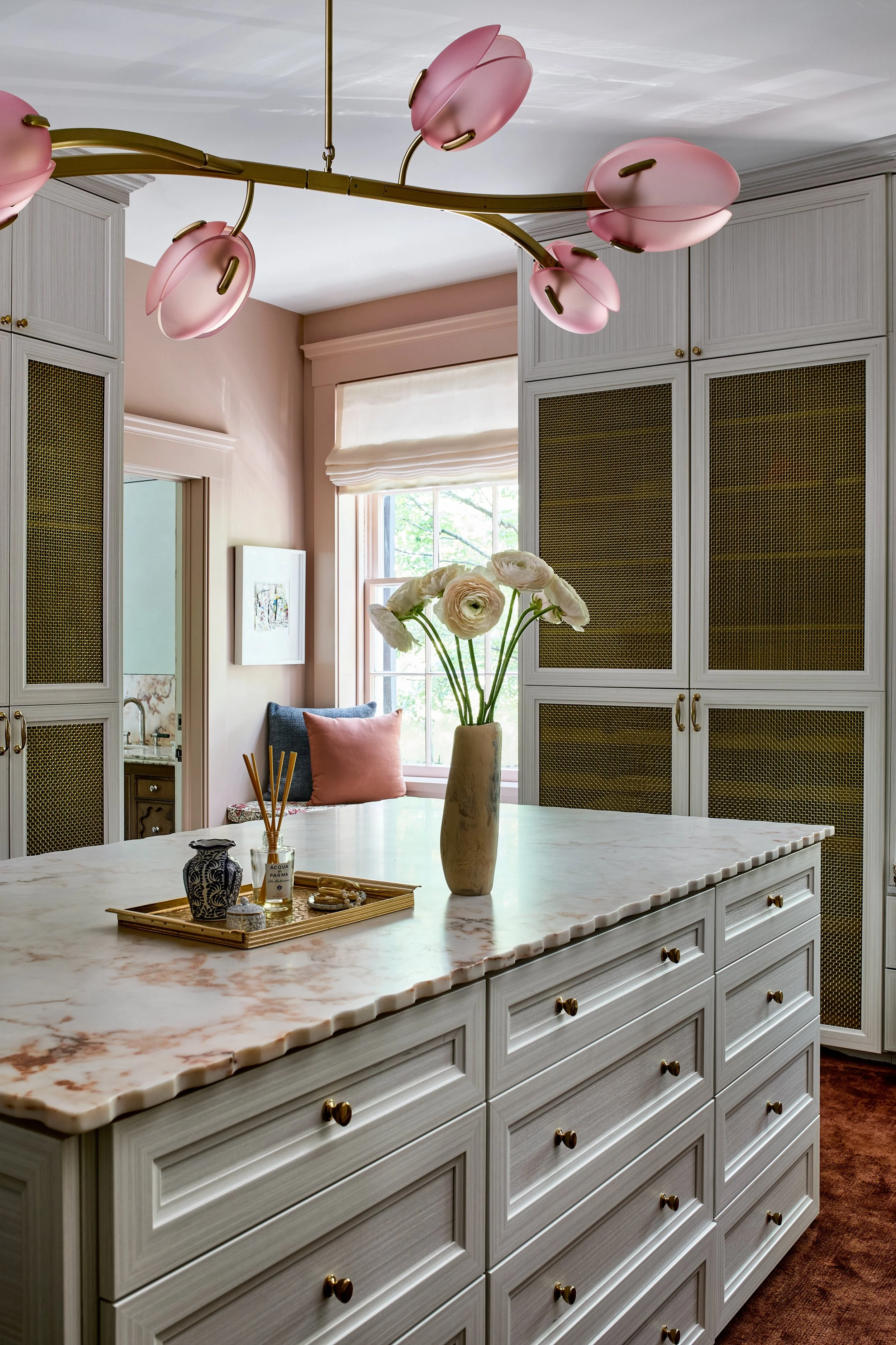 A kitchen island with a marble top, a vase with white flowers, and a tray with decor items, surrounded by white cabinets and a window with a view of trees.