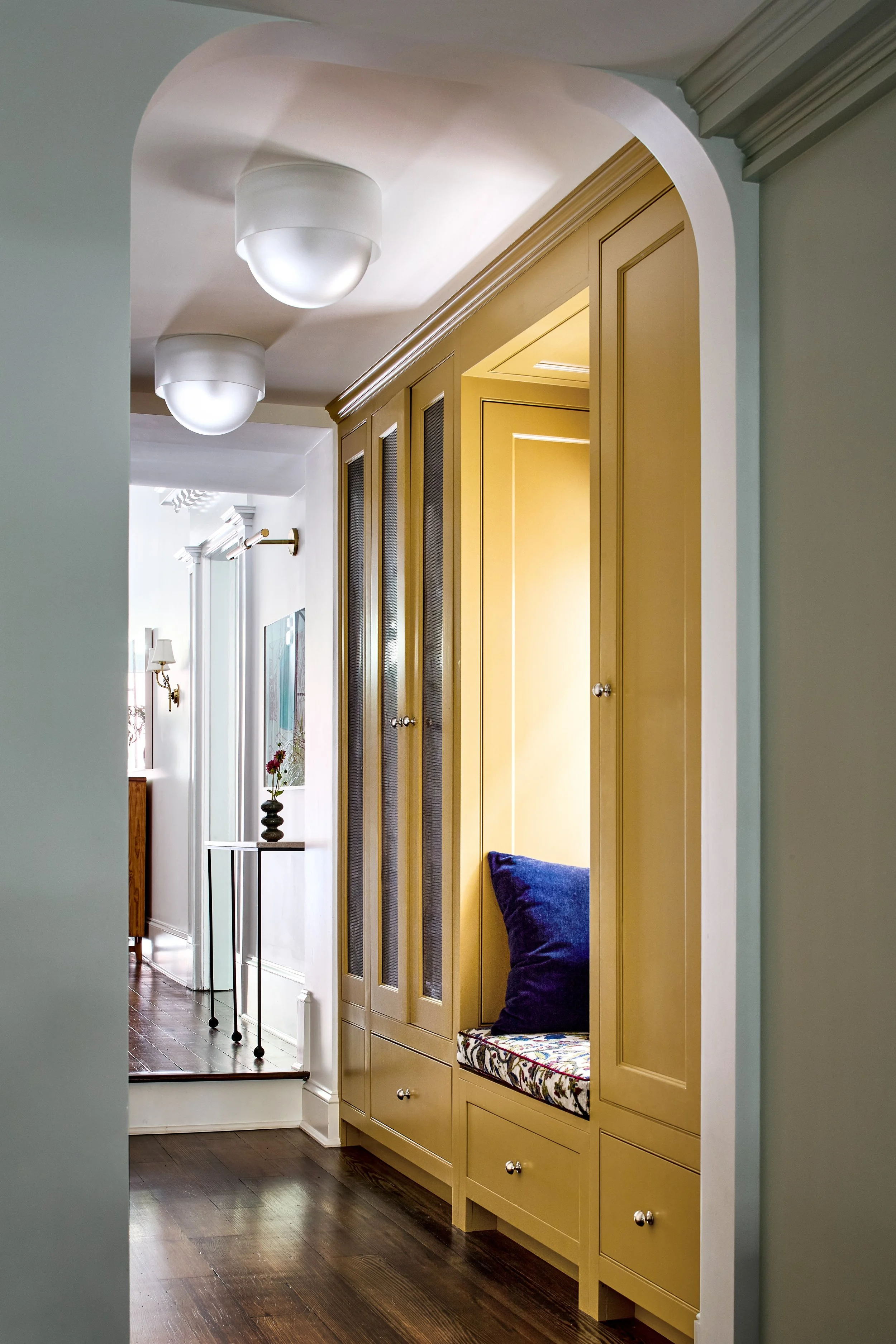 Interior view of a hallway with wooden flooring, leading to a built-in yellow cabinet with a seating nook and decorative cushions, under ceiling-mounted lights.