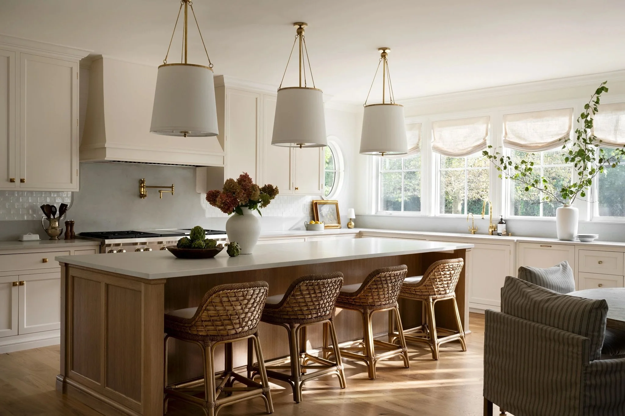 Bright kitchen with white cabinets, a large island with four woven chairs, pendant lights, and a window with roman shades. Decor includes a vase with flowers on the island and a large plant near the window.