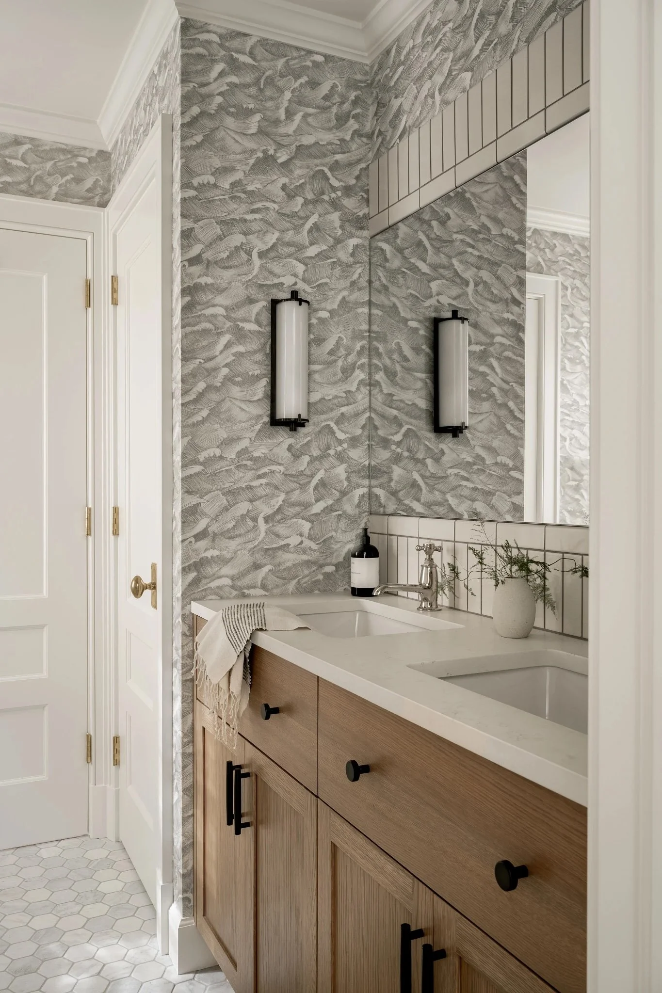 Bathroom vanity area with a white countertop, a wooden cabinet with black handles, a large mirror, gray patterned wallpaper, and two black-framed vertical wall-mounted lights.