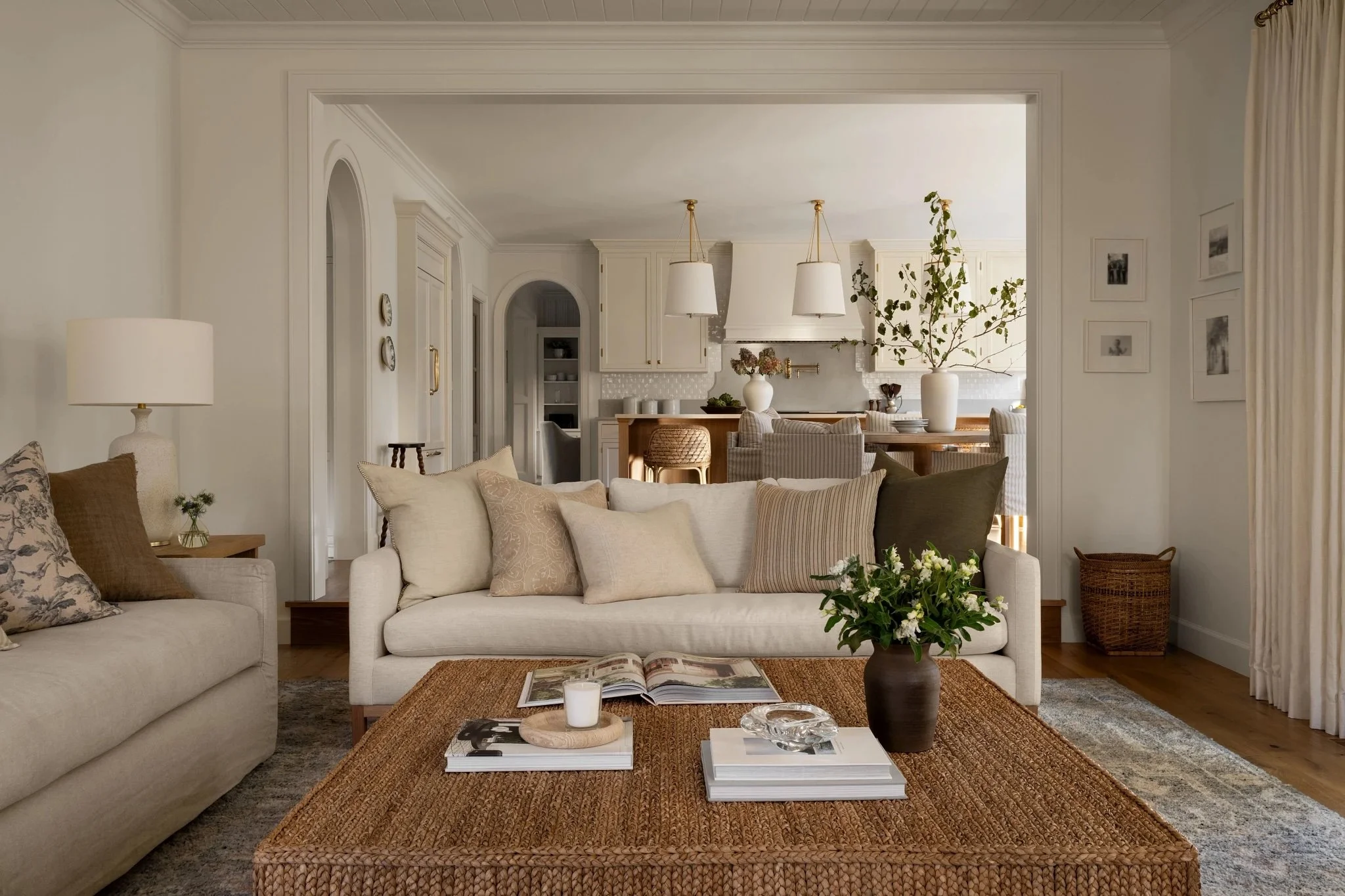 Living room with beige sofa, assorted pillows, wooden coffee table with books, flowers, and candles, and a dining area in the background with a large vase and greenery.