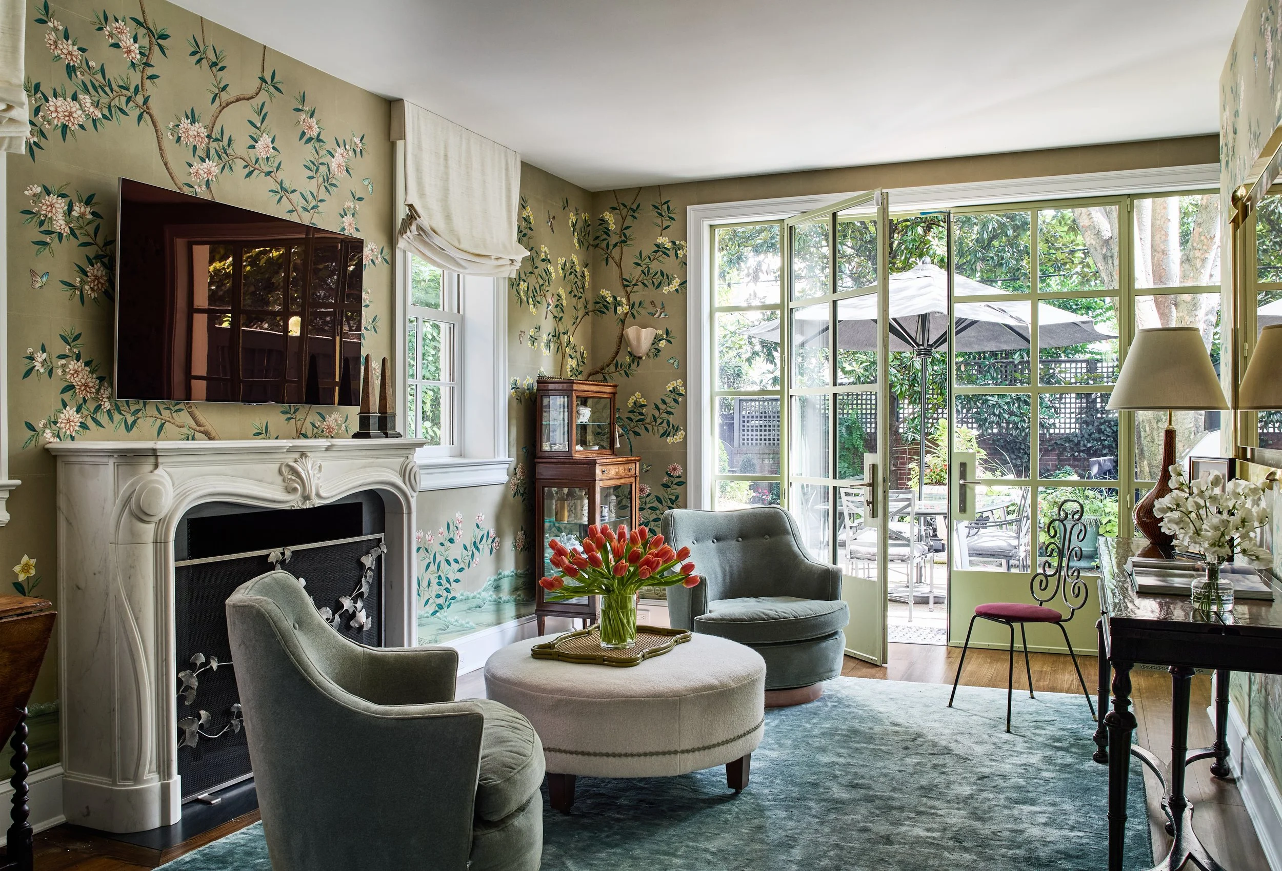 Living room with green floral wallpaper, a fireplace, a flat-screen TV, two armchairs, a round ottoman with a vase of red tulips, and an open door leading to a patio with outdoor furniture and an umbrella.