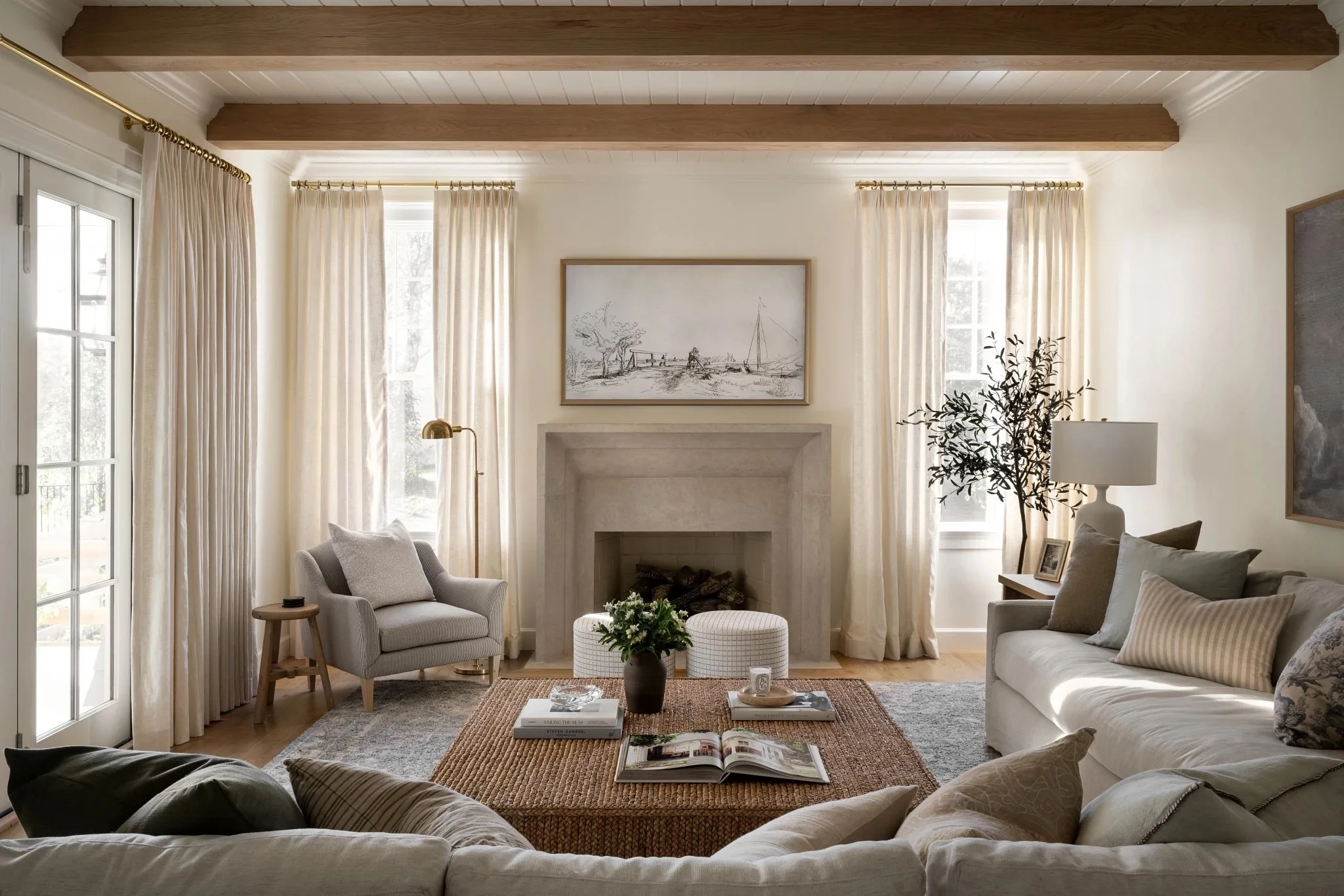 Living room with cream-colored sofa, armchair, stone fireplace, and wooden beams on ceiling. Windows with cream curtains, table with flower vase, and framed artwork above fireplace.