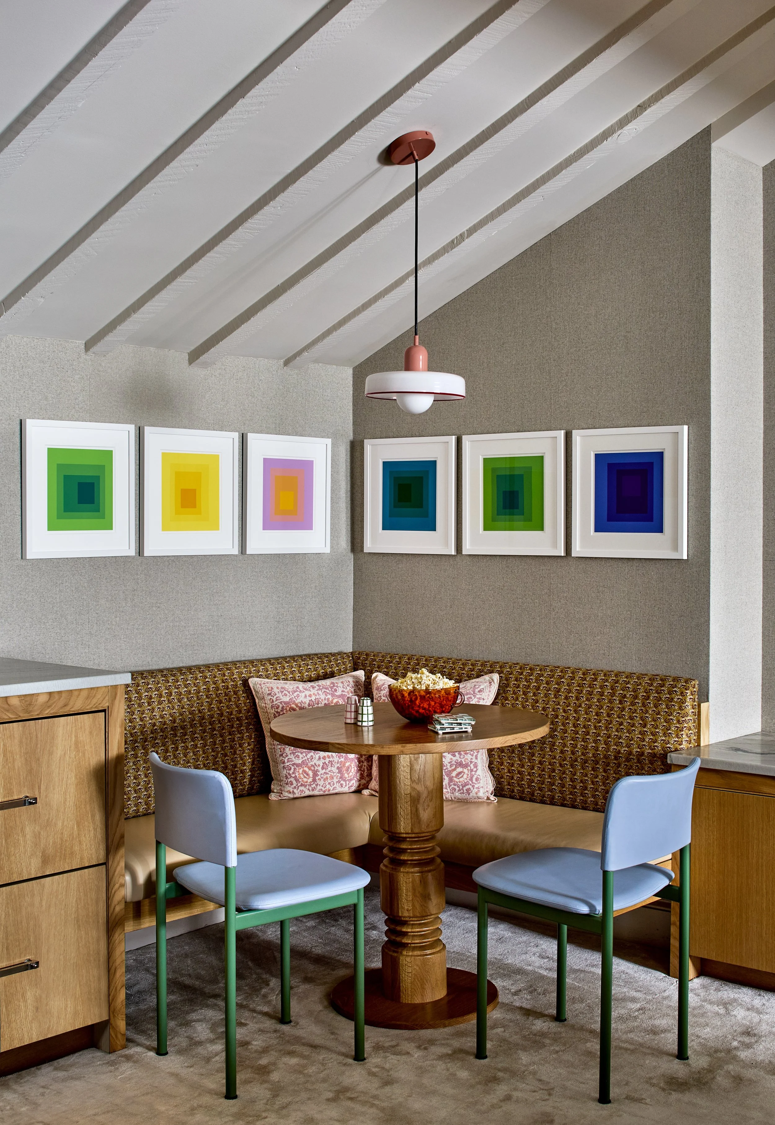 A cozy dining nook with a round wooden table, two light blue chairs, a built-in corner bench with patterned cushions, colorful framed art prints on the wall, and a modern hanging pendant light.