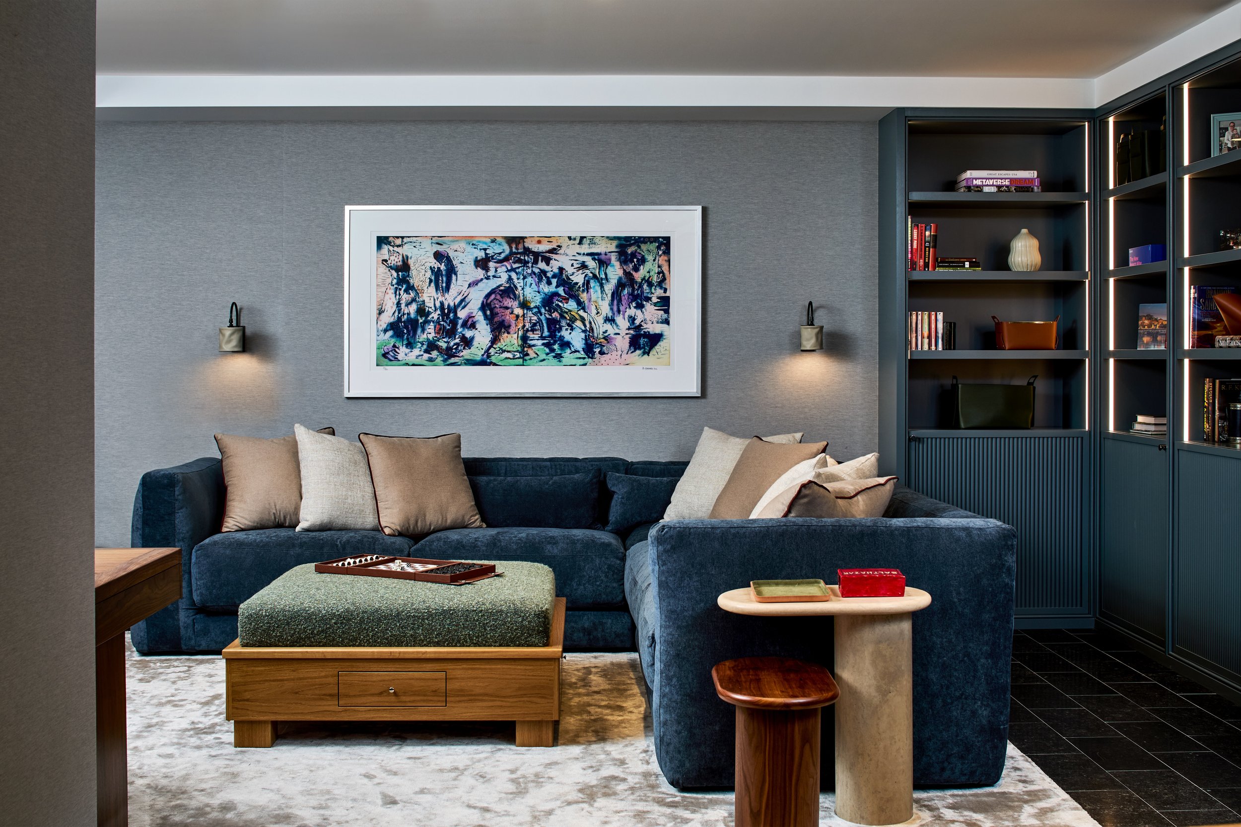 Modern living room with blue sectional sofa, beige and white pillows, abstract artwork on gray wall, built-in dark blue bookshelves, side table with decorative boxes, and a textured ottoman on a light-colored rug.