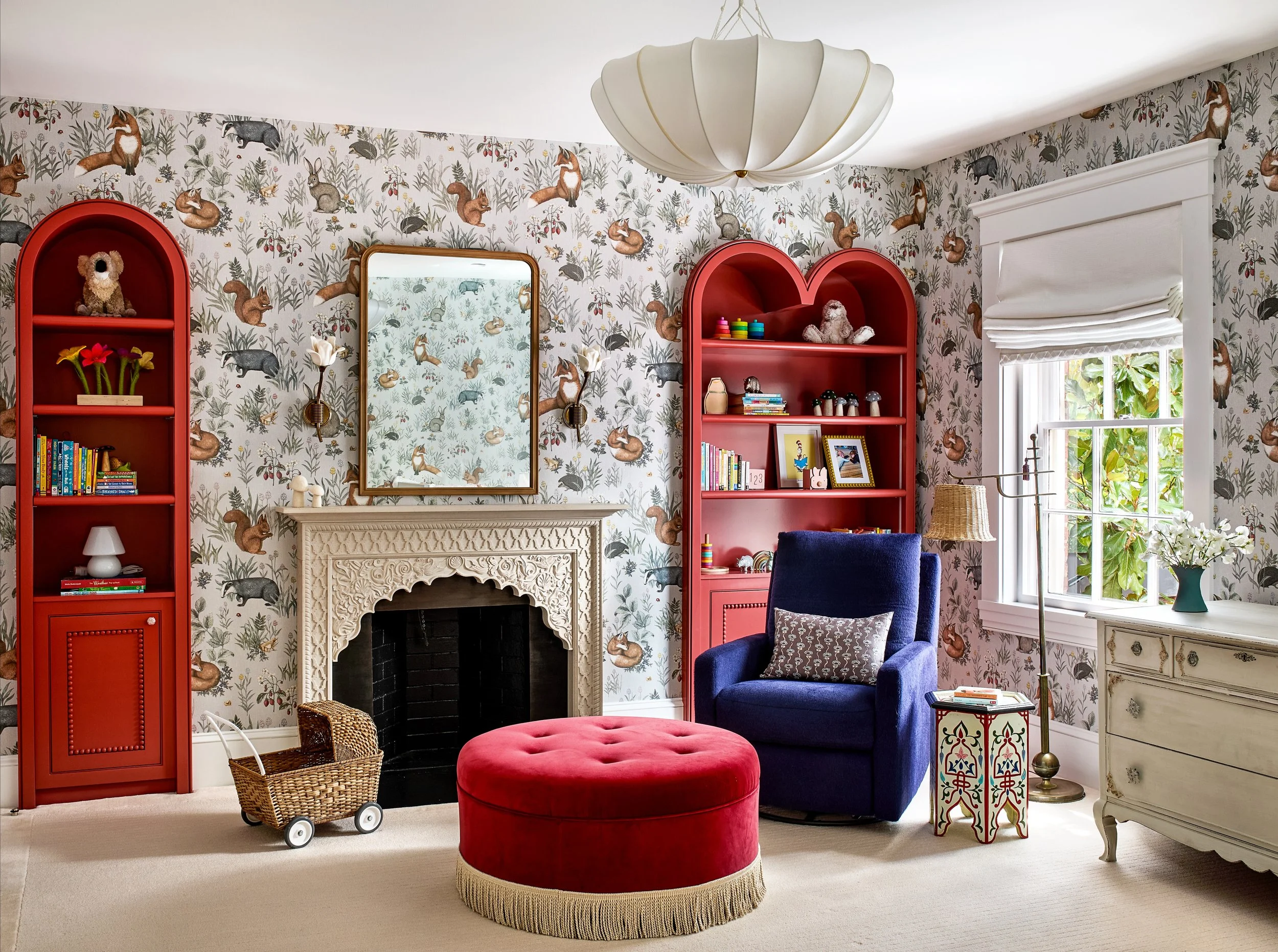A brightly decorated living room with whimsical wallpaper featuring animals, red arched bookshelves, a white ornate fireplace, a navy blue armchair with a patterned pillow, a round red ottoman, a wicker wagon, a white dresser, and a window with white blinds and a vase of white flowers.