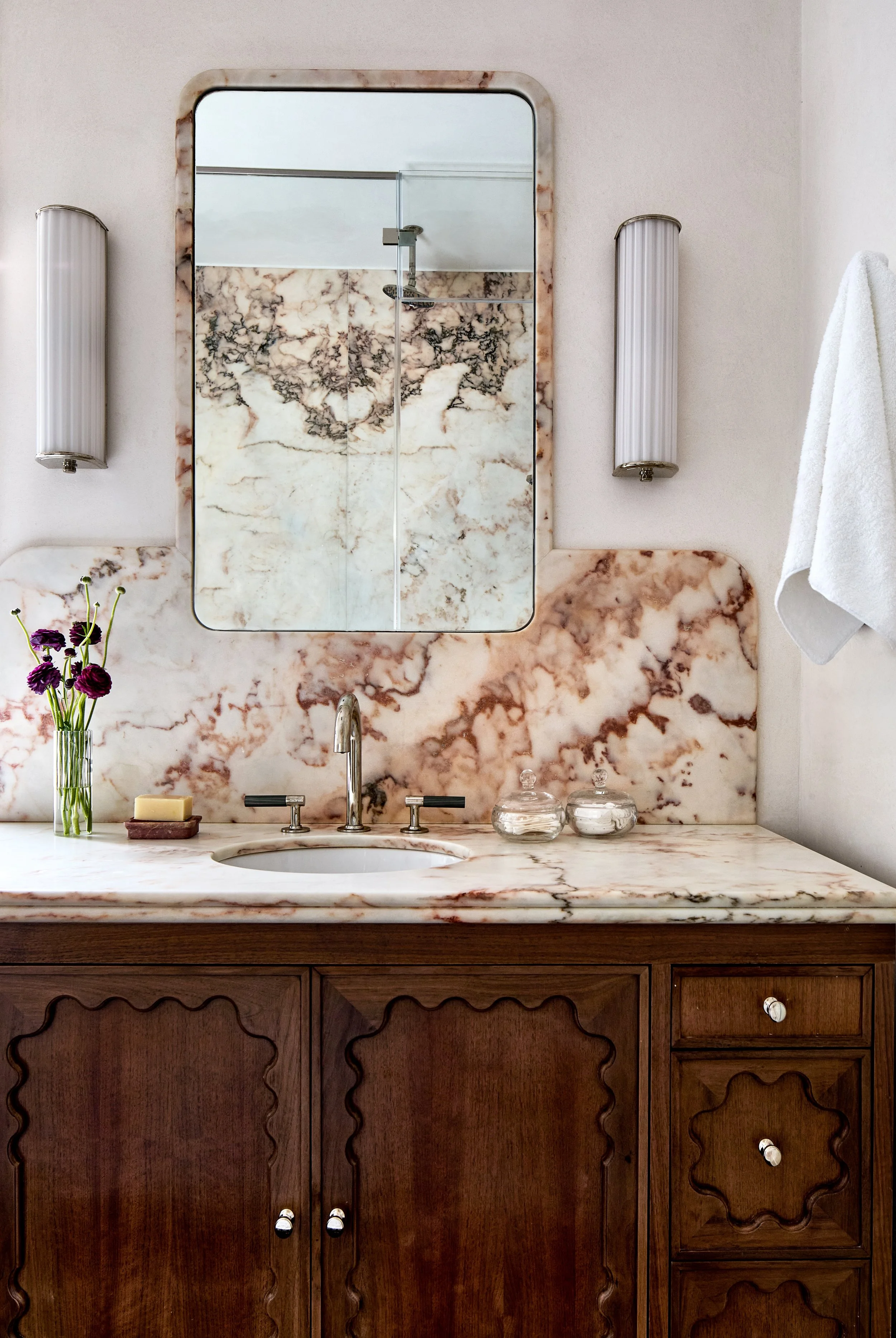 Bathroom vanity with marble countertop and backsplash, a mirror, two wall-mounted lights, a white towel hanging on the wall, a vase with purple flowers, two jars, a bar of soap, and a tiled shower area reflected in the mirror.