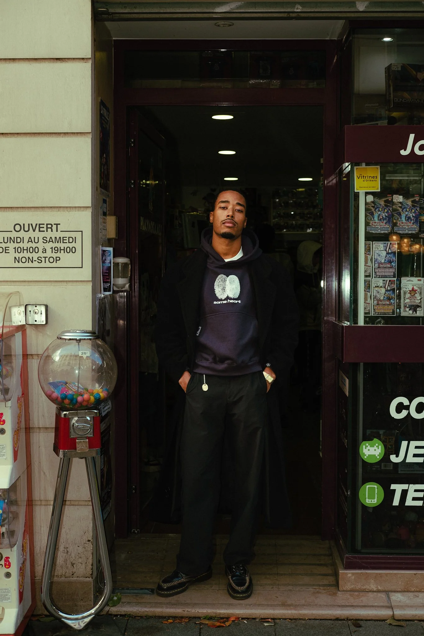 Jeune homme debout devant une boutique, portant un sweat à capuche noir et un pantalon noir, mains dans les poches, regard sérieux.