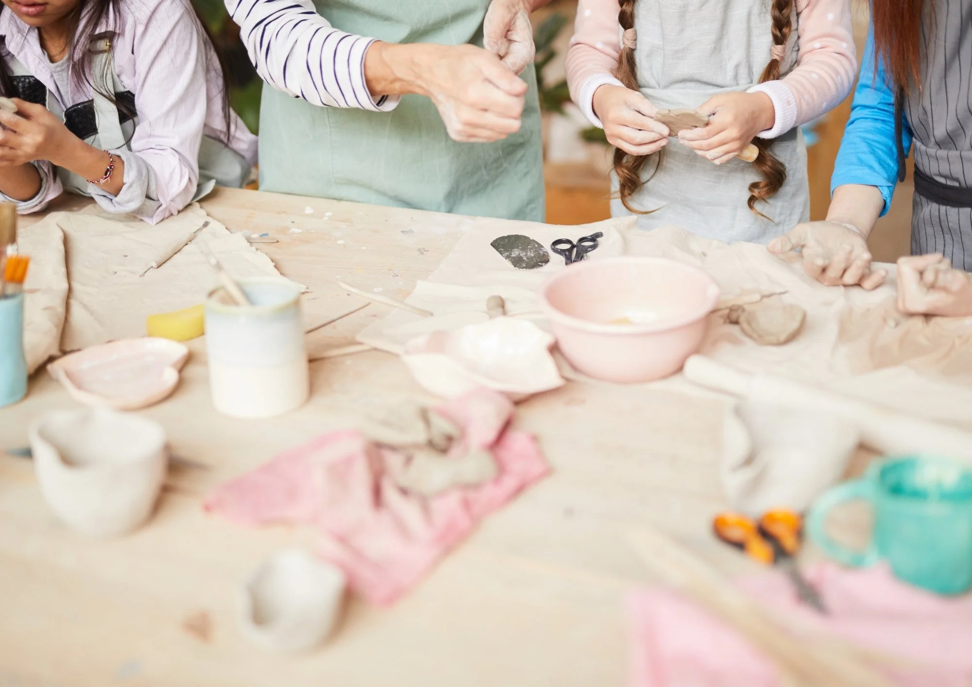 Groupe de personnes en train de créer de la poterie avec de l'argile sur une table en bois, entourées d'outils et de pièces de céramique.