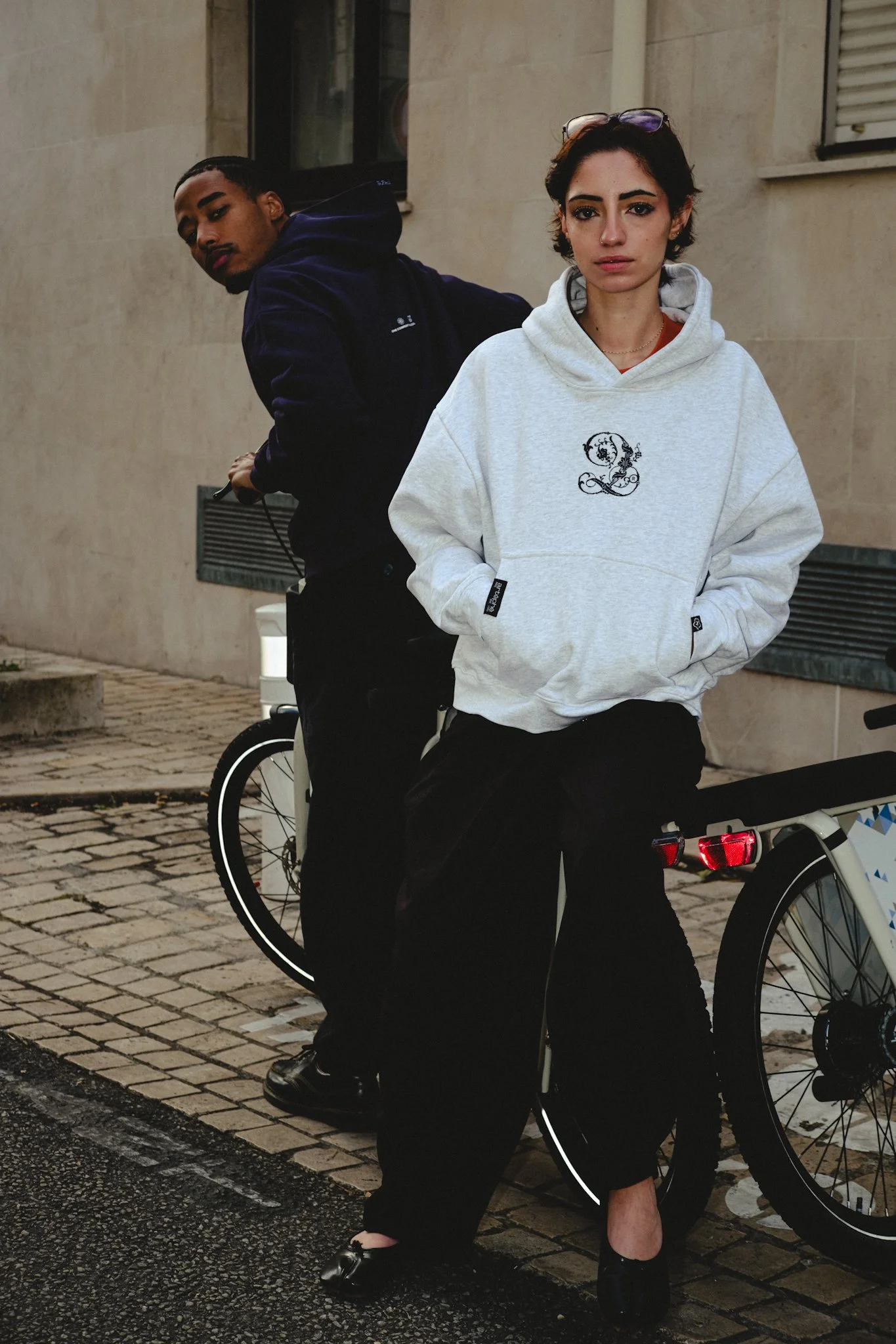 Une femme et un homme posant sur un trottoir en ville, avec des vélos à côté, la femme porte un sweat à capuche gris clair, et l'homme porte un sweat ou une veste foncée.