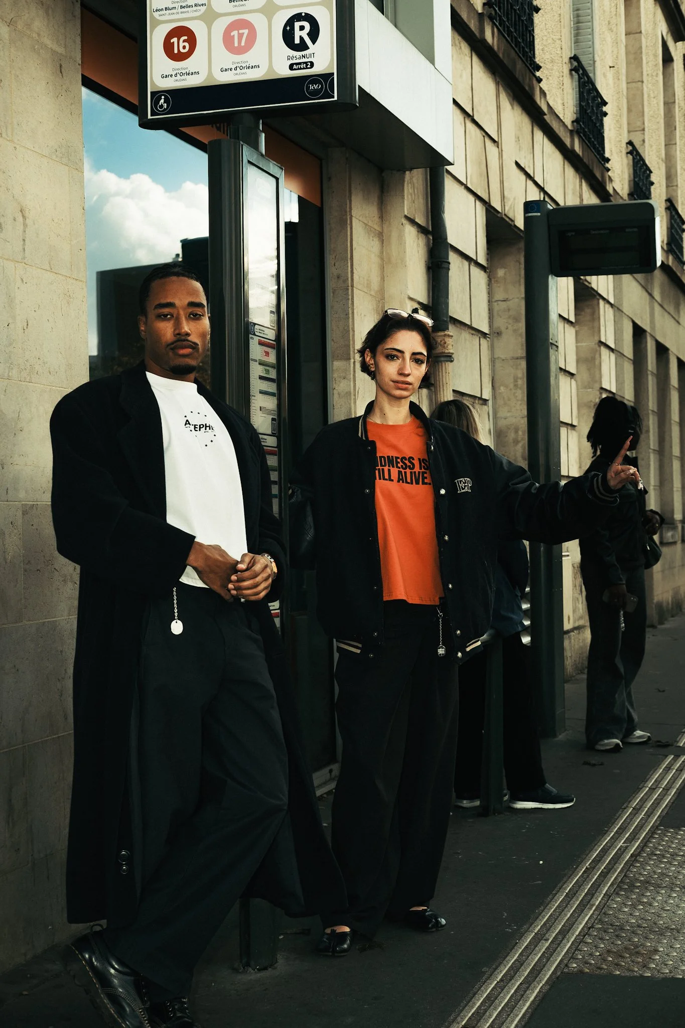 Deux personnes debout à une station de bus ou de métro, l'une à gauche portant une veste noire et un pantalon noir, l'autre à droite portant une veste noire et un t-shirt orange avec un message en anglais. D'autres personnes en arrière-plan et un panneau d'informations de transit au-dessus.
