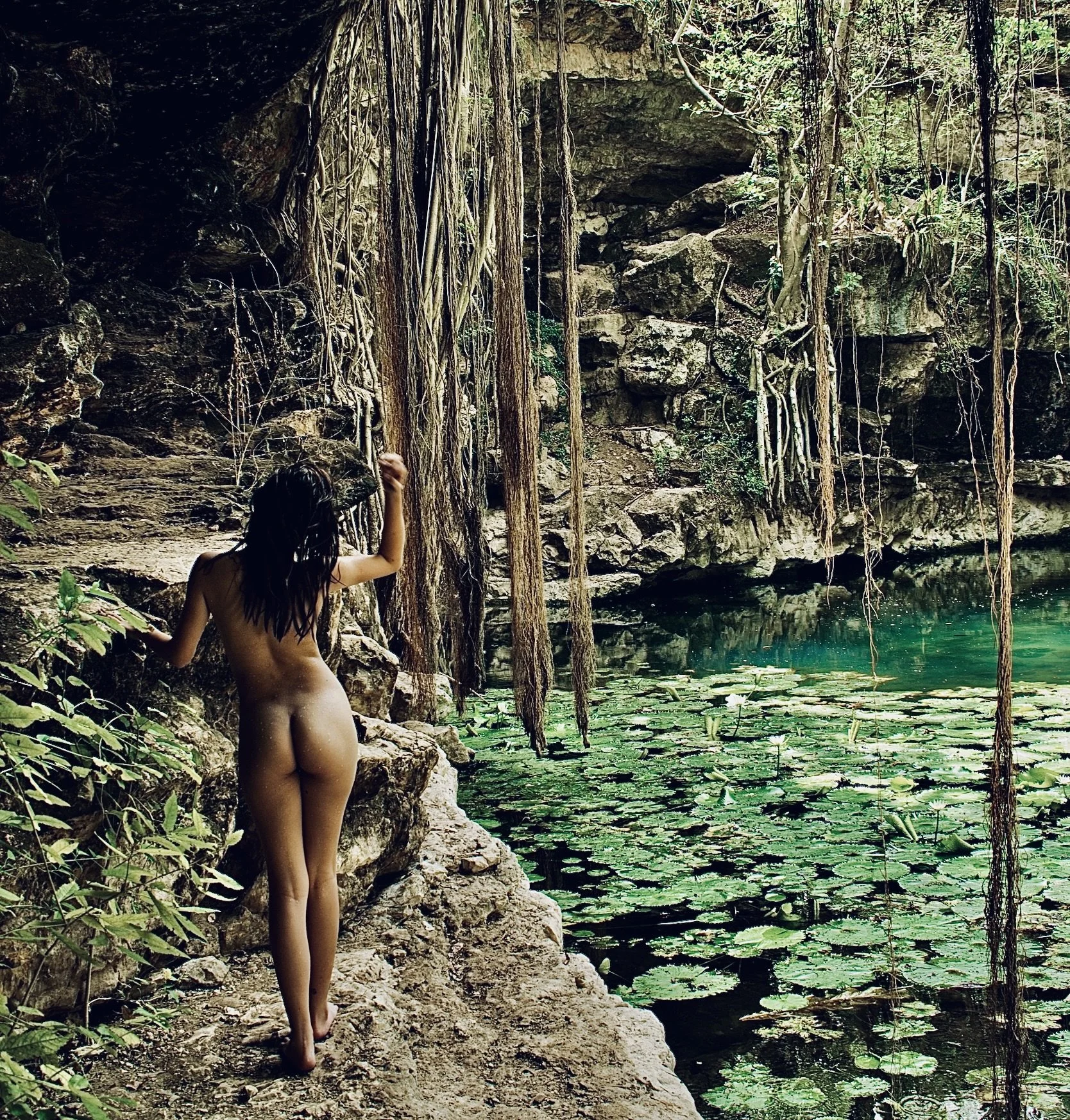 Persona junto a un cenote rodeado de vegetación y rocas, con agua turquesa y plantas acuáticas.