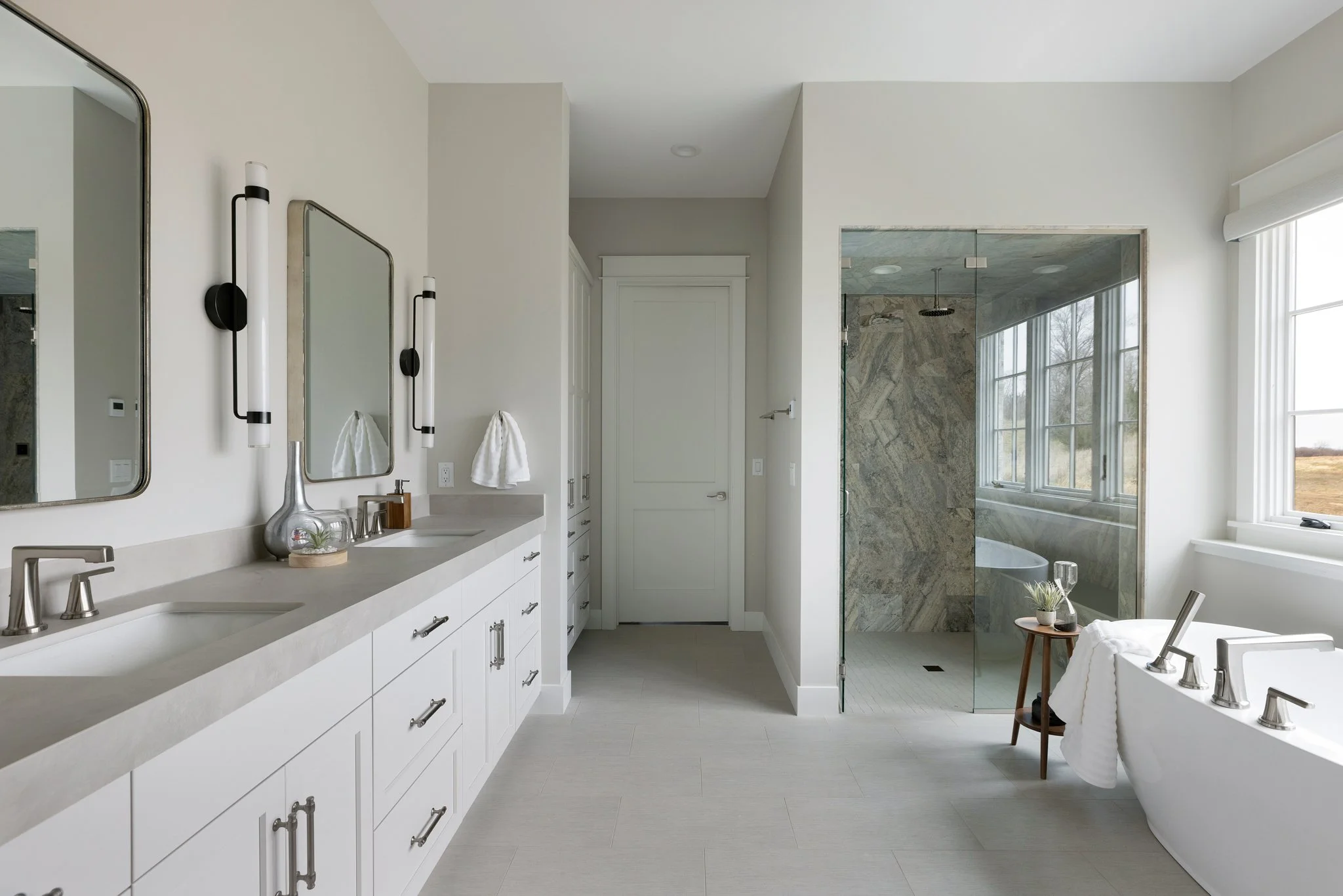 Modern bathroom with double vanity, large mirror, wall sconces, white cabinets, enclosed glass shower with marble tiles, and a freestanding bathtub, illuminated by natural light from large windows.