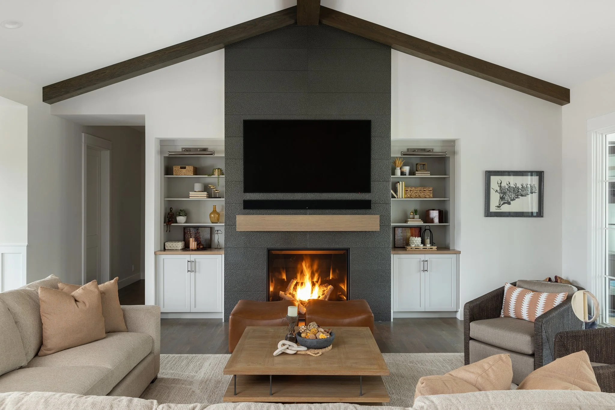 Living room with a modern fireplace, mounted TV above, beige sofa, and gray armchair with striped pillow, natural wood coffee table, built-in shelves with decor, and a window on the right.
