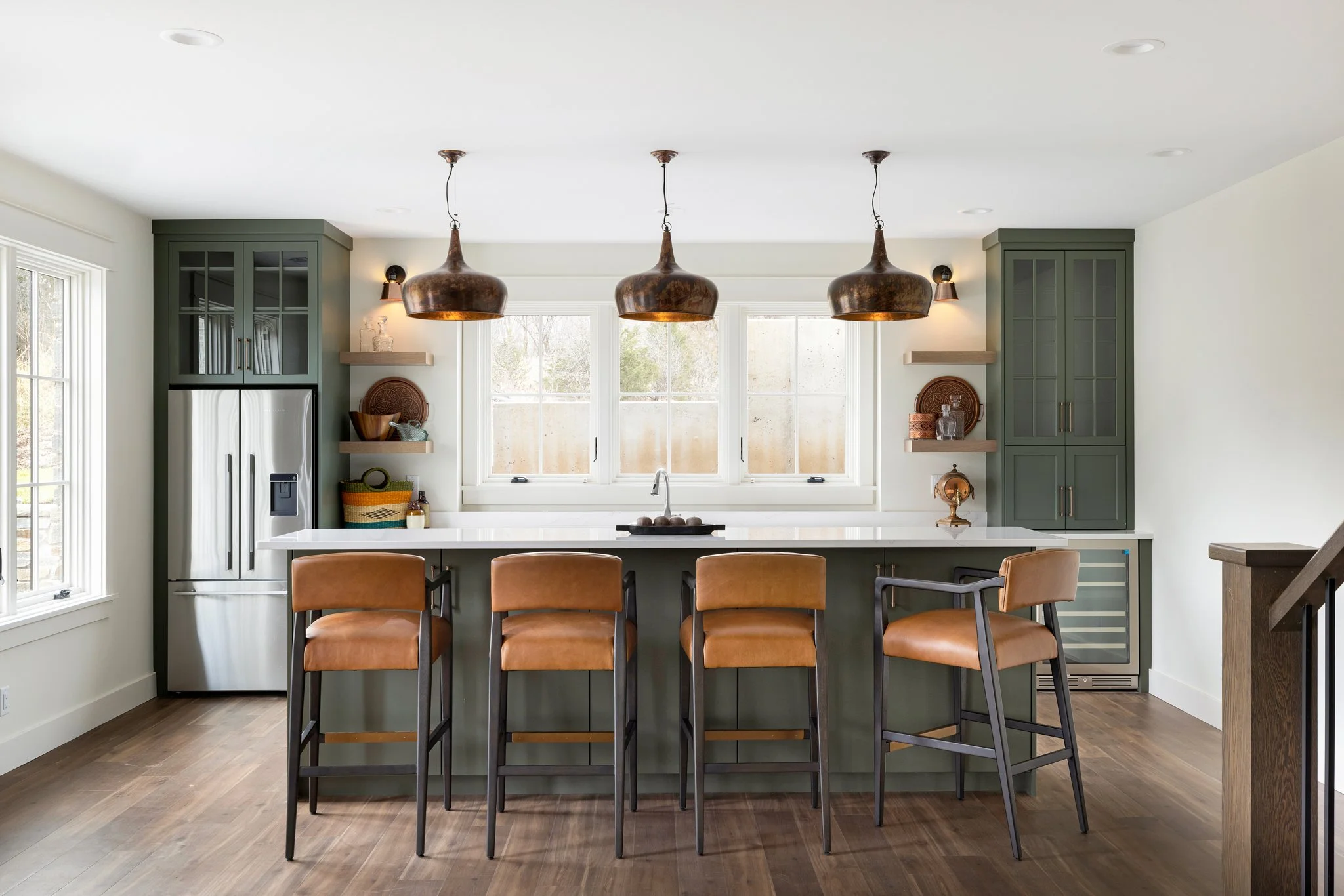Modern kitchen with green cabinets, white countertops, four brown leather chairs at the island, three copper pendant lights, and a large window behind the sink.