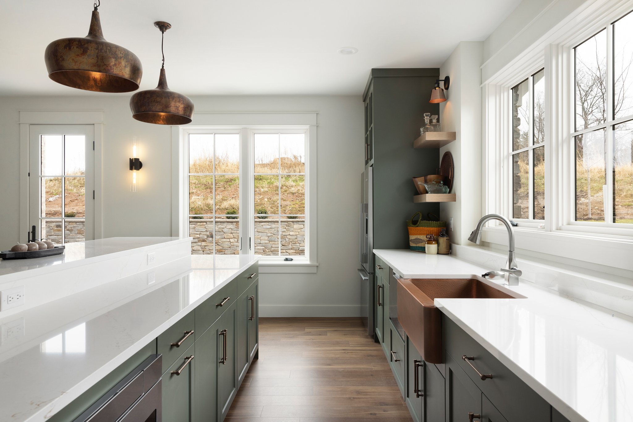 Modern kitchen with white countertops, green cabinets, copper sink, large windows, wood flooring, and pendant lights.
