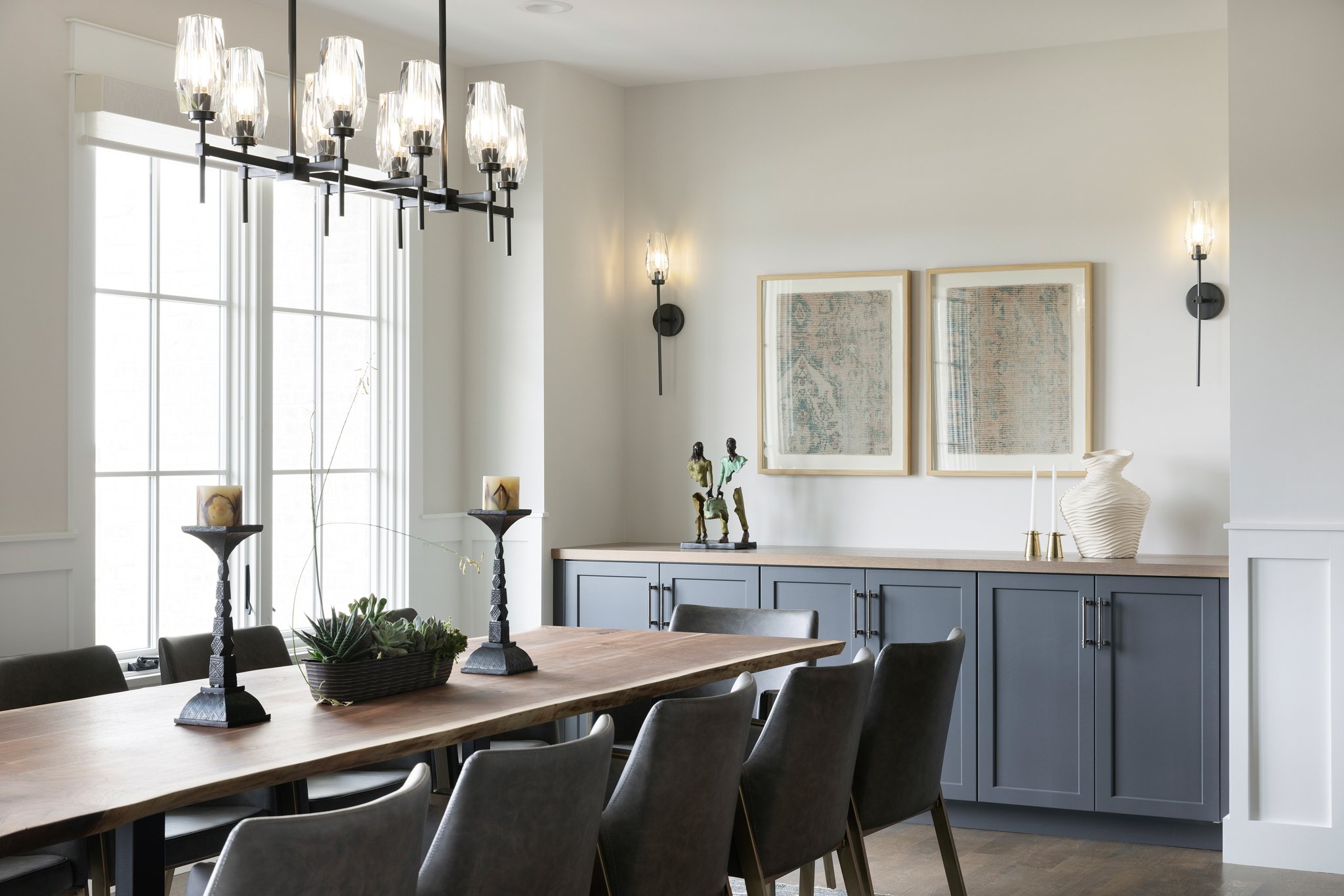 Modern dining room with a wooden table, dark chairs, large window, black chandelier, and a blue sideboard with art and decorative items.