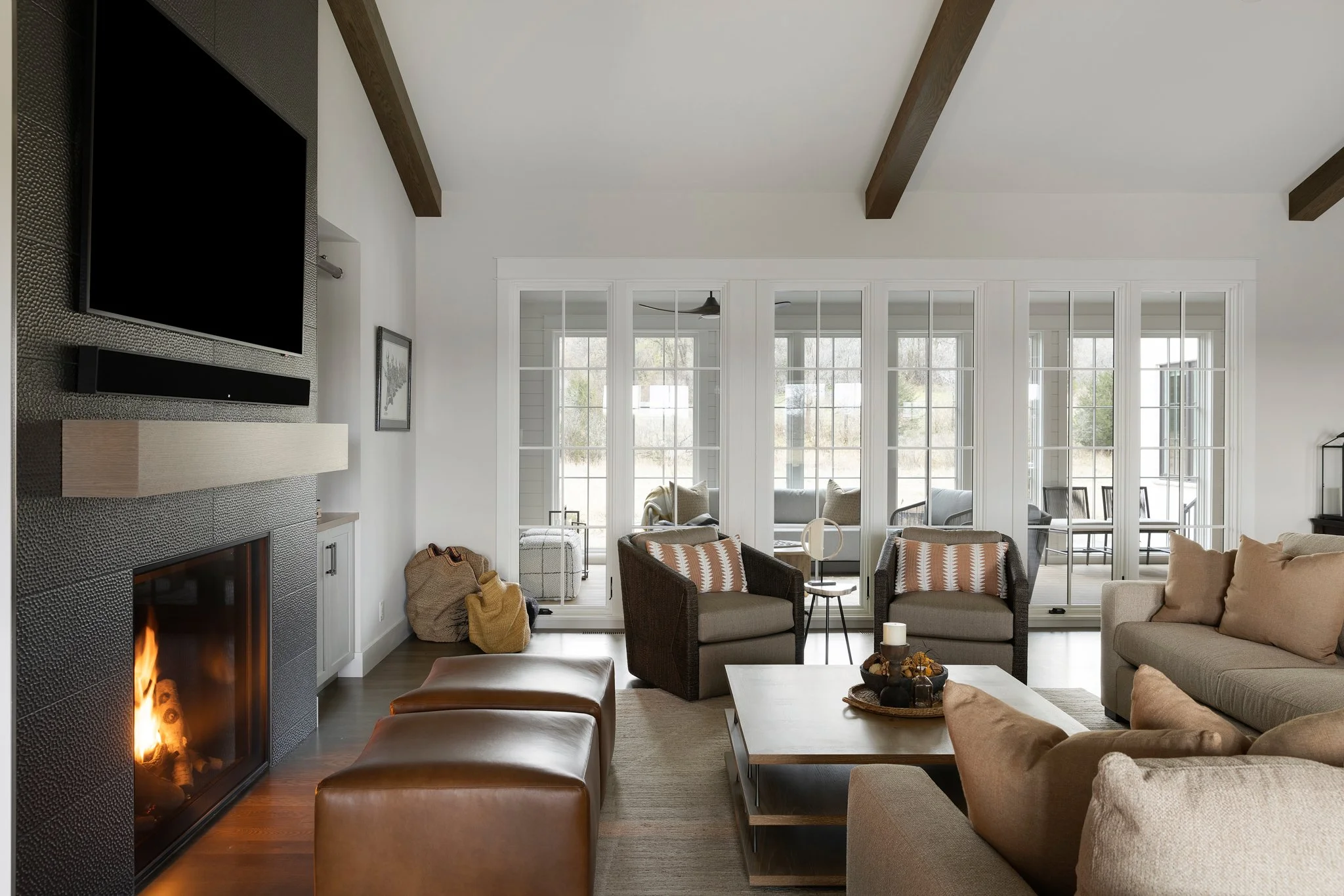 Living room with a fireplace, beige and brown furniture, large windows with white frames, and a view of a porch with chairs outside.