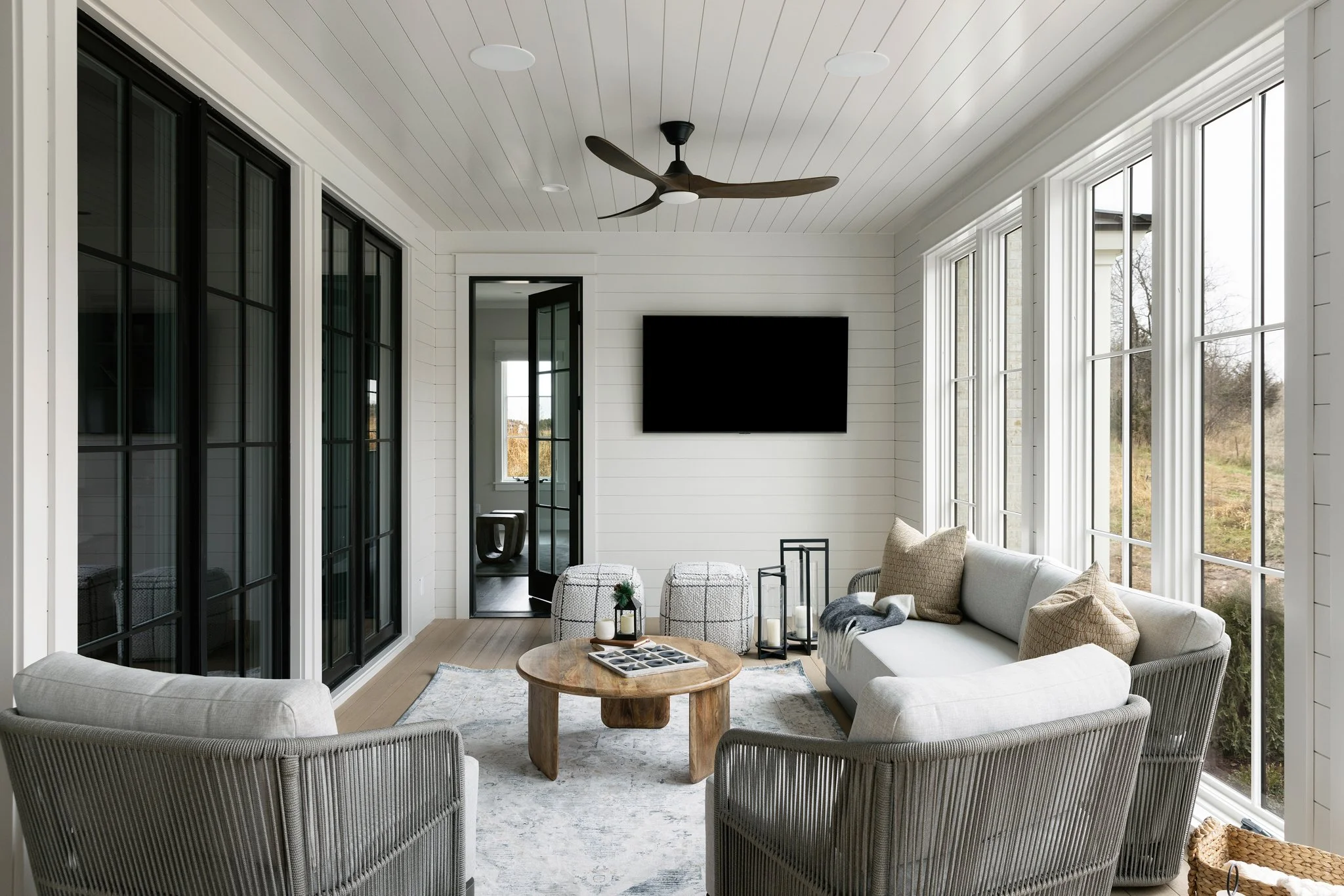 Sunroom with white shiplap walls, large windows, a wall-mounted black TV, gray cushioned seating, a round wooden coffee table, decorative lanterns, and a ceiling fan.